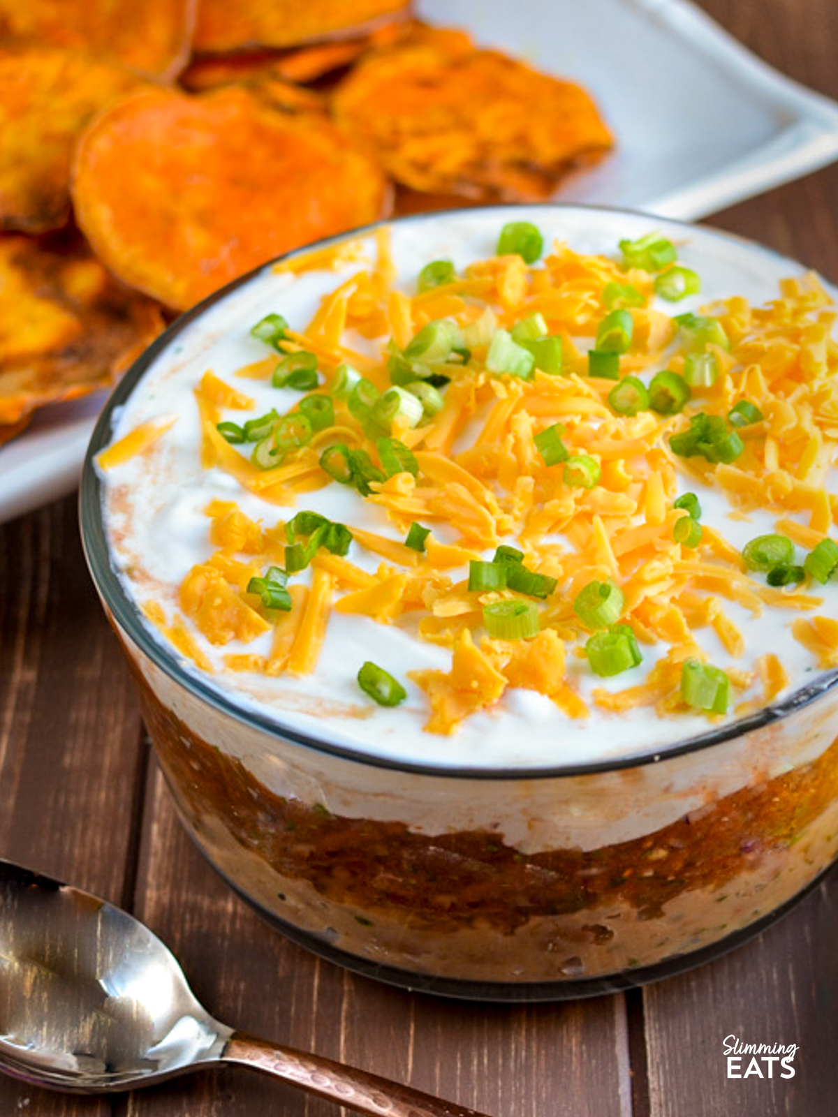mexican layered dip in a bowl next to a yellow, orange, white and brown grey towel and a plate of homemade sweet potato chips