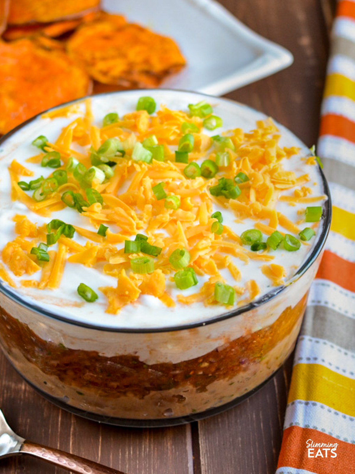 mexican layered dip in a bowl next to a yellow, orange, white and brown grey towel and a plate of homemade sweet potato chips