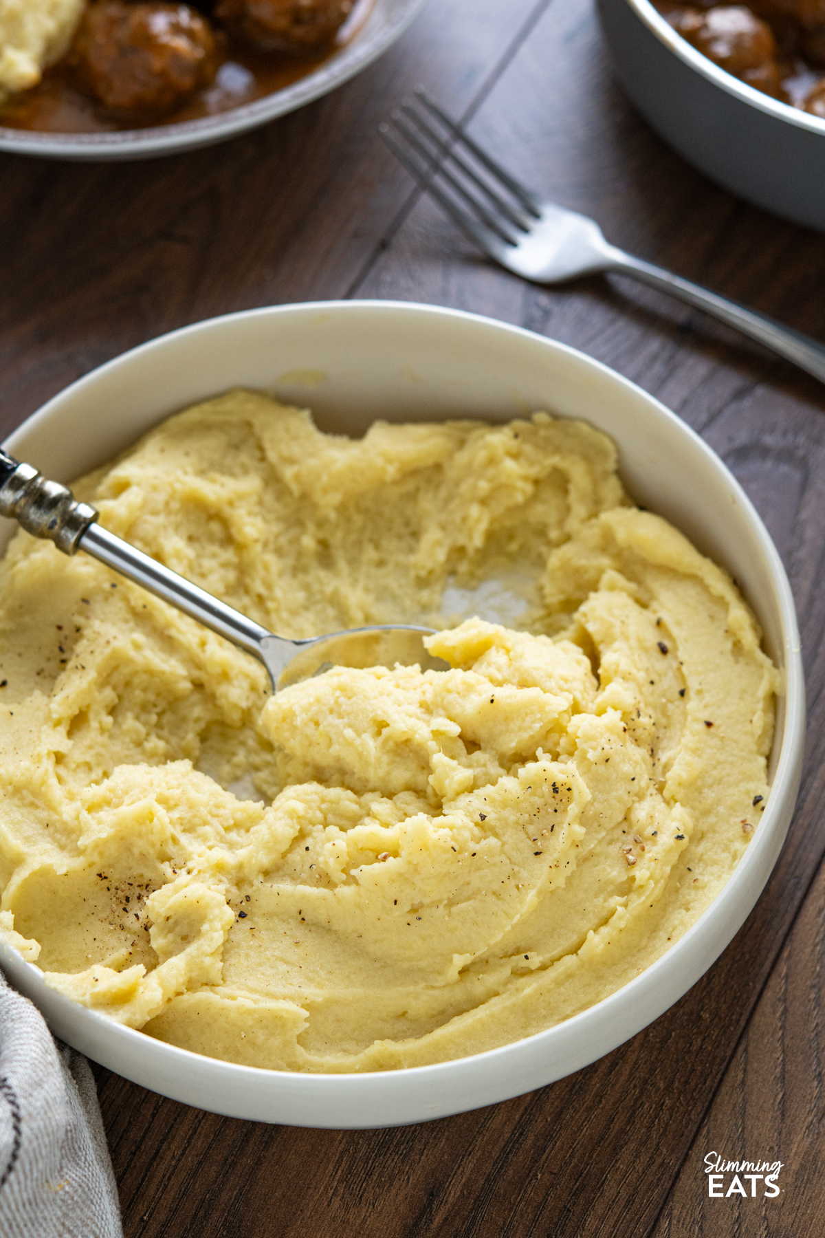 lighter mashed potatoes in a white bowl next to a pan with meatballs in gravy and a small bowl of green peas