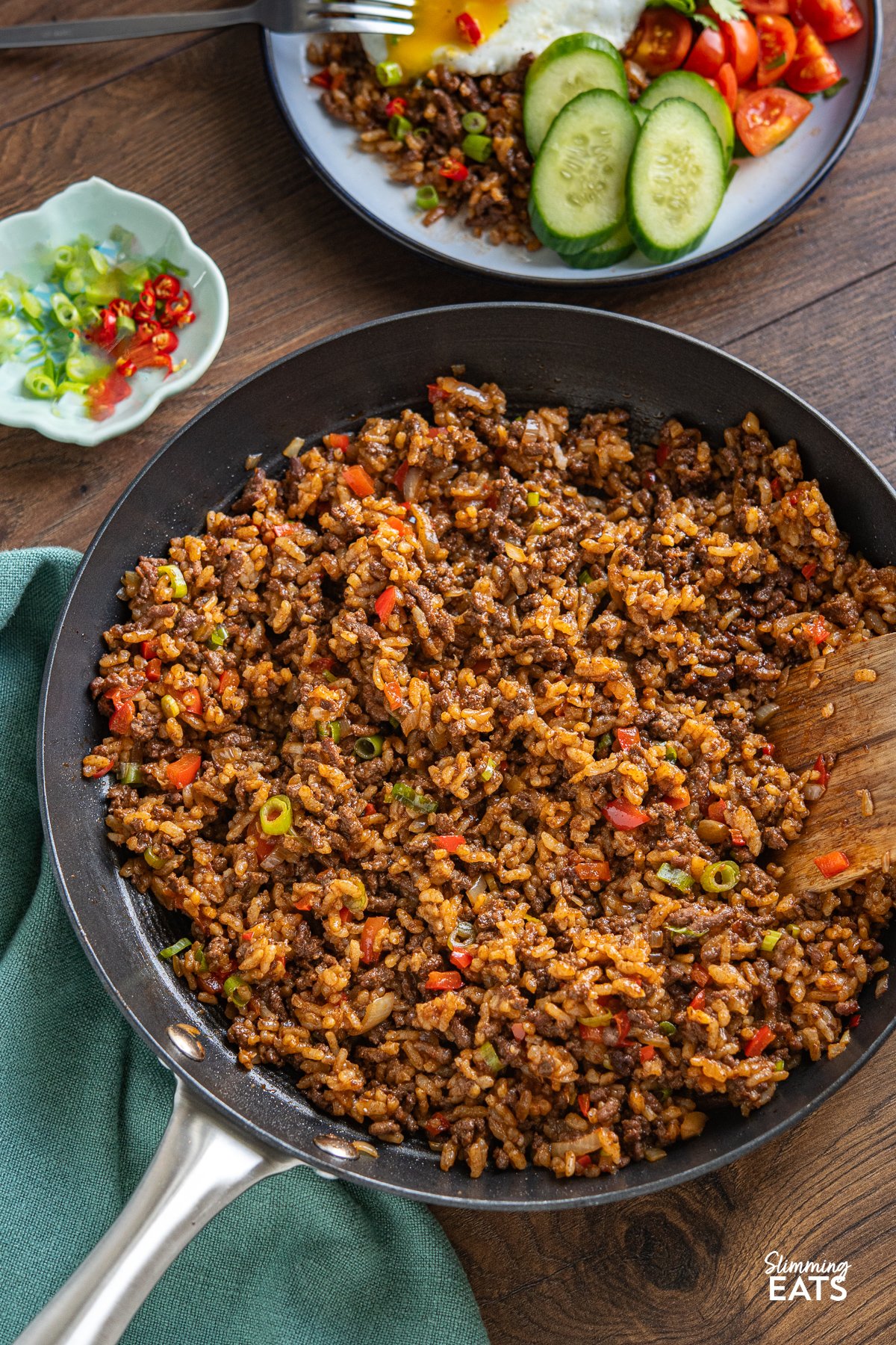 Beef Nasi Goreng in a black frying pan with wooden spatula, bowl of served up nasi goreng above and chilli and spring onions