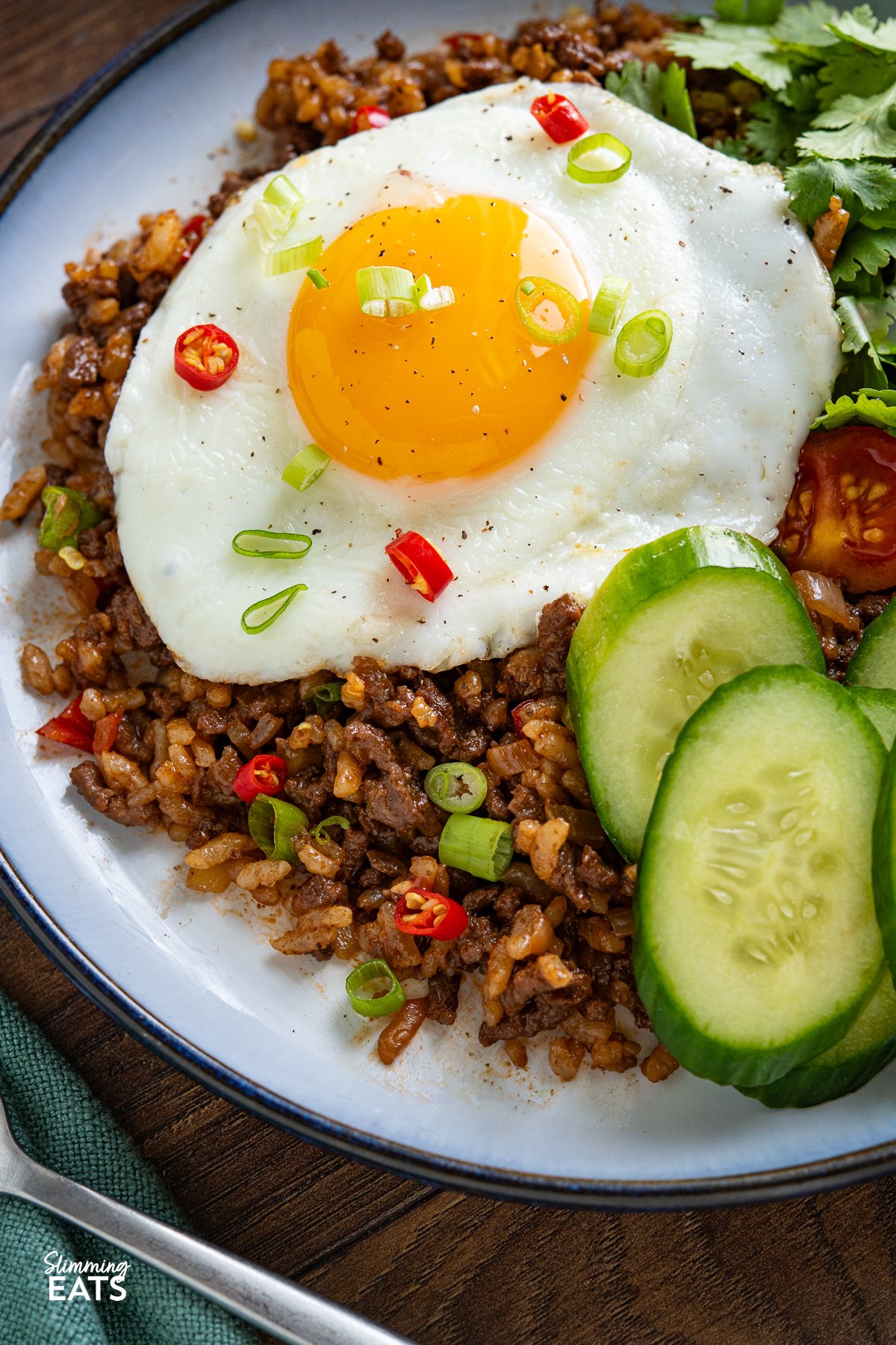 Discover the rich and savoury world of Indonesian cuisine with this tantalizing Nasi Goreng, a flavorful fried rice delight featuring beef mince, enhanced by a sweet and umami-packed soy sauce with a hint of spice. close up of Served up nasi goreng topped with fried egg and served with tomatoes and cucumber in a pale blue bowl with navy rim.