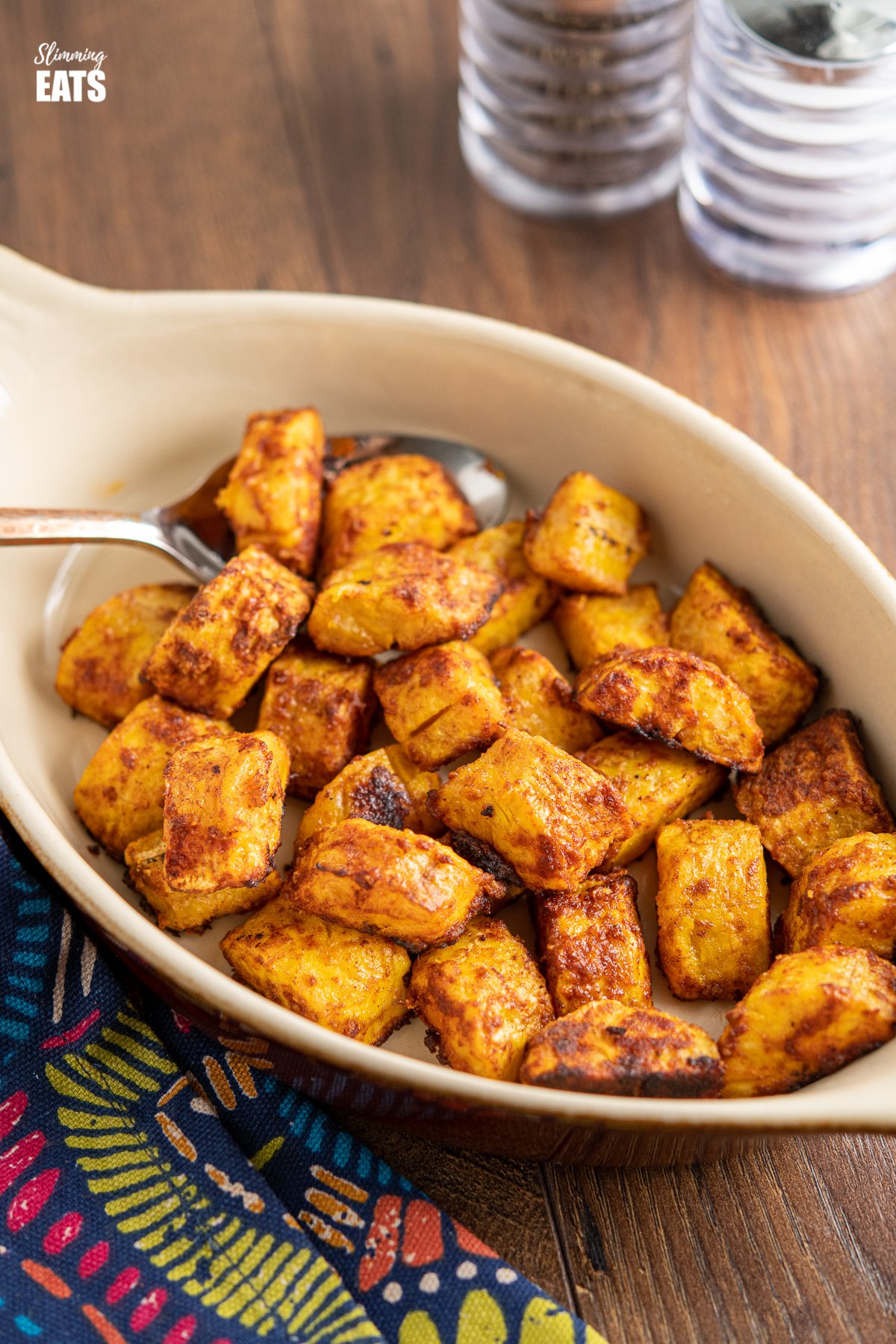 baked sweet plantain in a oven proof brown dish with spoon on wooden board