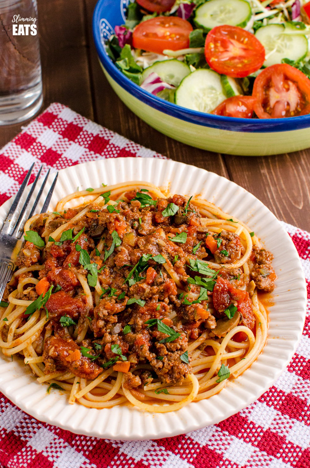 bolognese on spaghetti pasta on white ridged plate on red checked fabric with salad and water in background