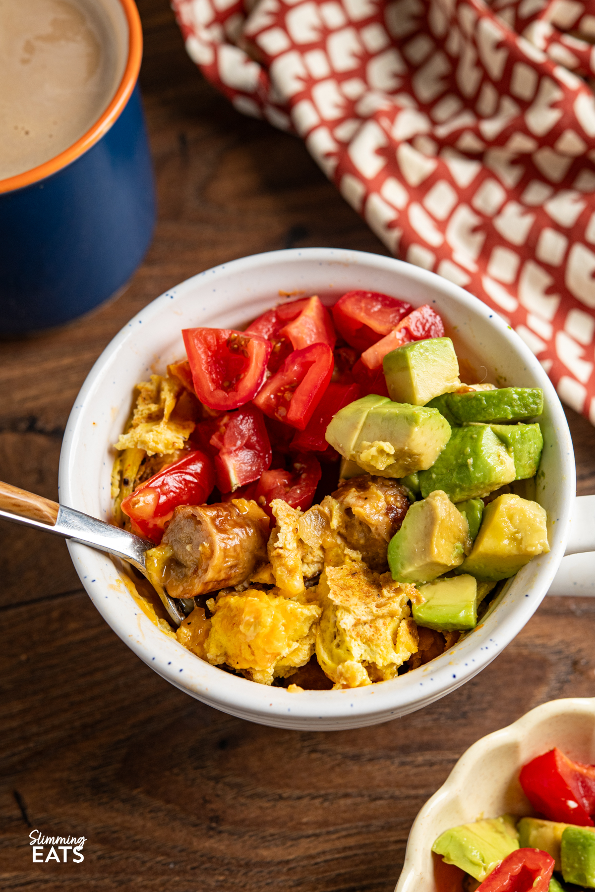Need a quick breakfast that still feels filling? This Breakfast in a Mug is perfect for those mornings when you want something warm and satisfying, without spending ages in the kitchen. close up of fork in Breakfast in a Mug topped with tomatoes and avocado and blue mug with coffee and brown and cream towel above.