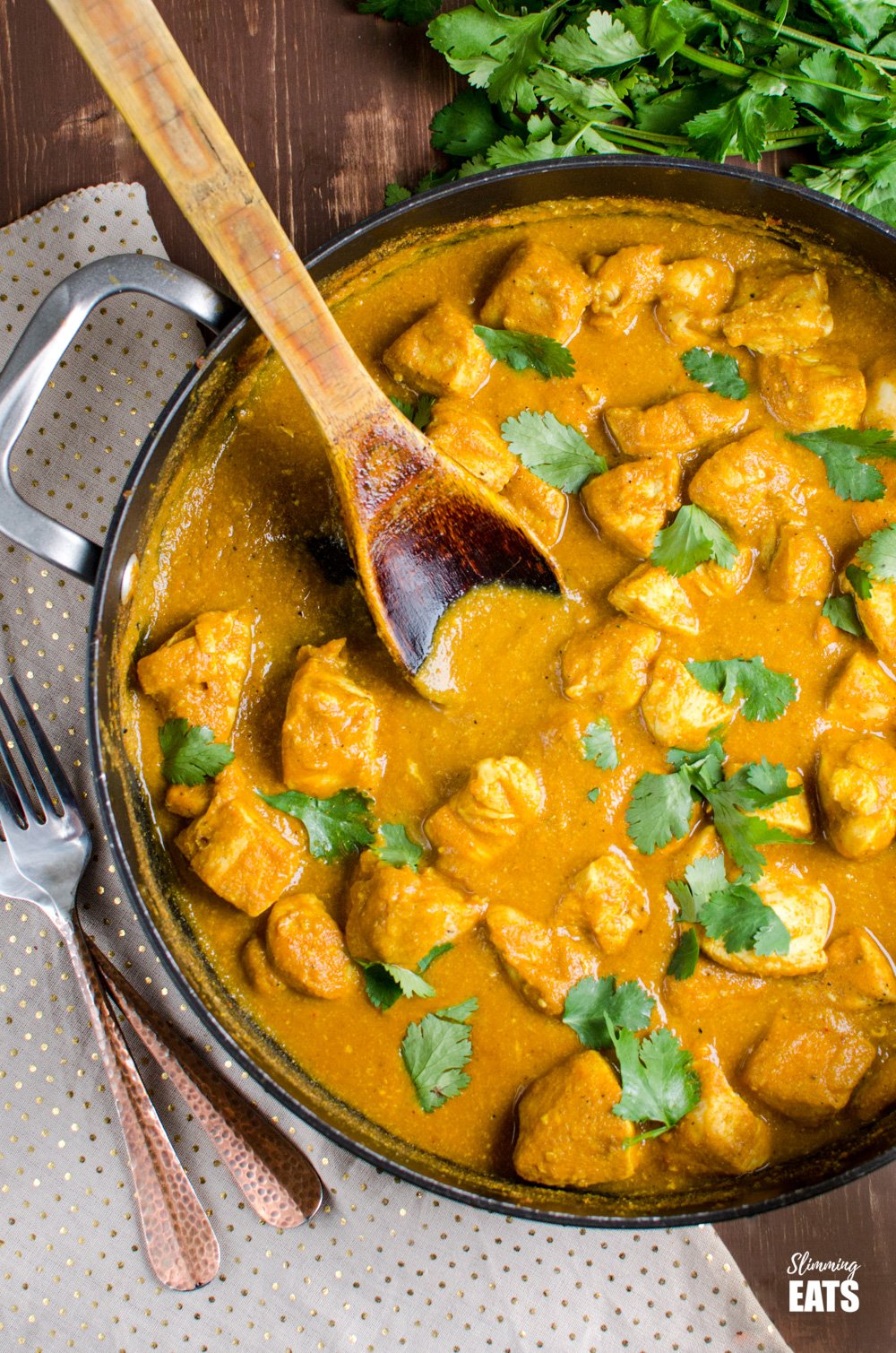 Chicken Korma in a deep black frying pan with wooden spoon, scattered with fresh coriander on wooden board with two forks