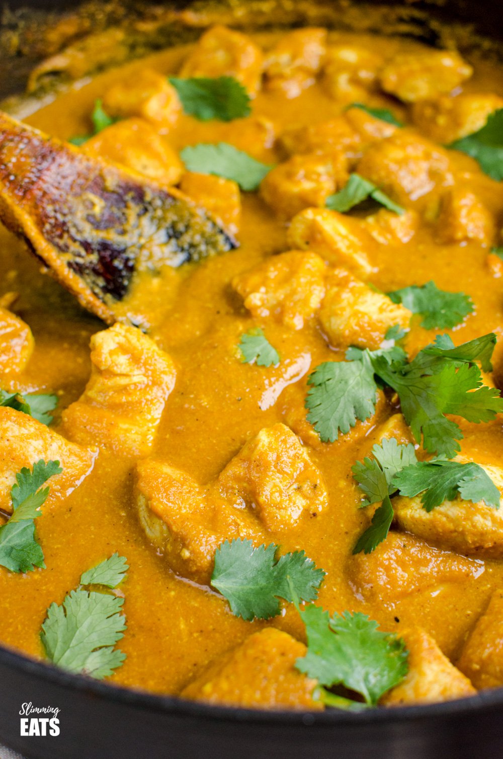 close up of Chicken Korma in a deep black frying pan with wooden spoon, scattered with fresh coriander