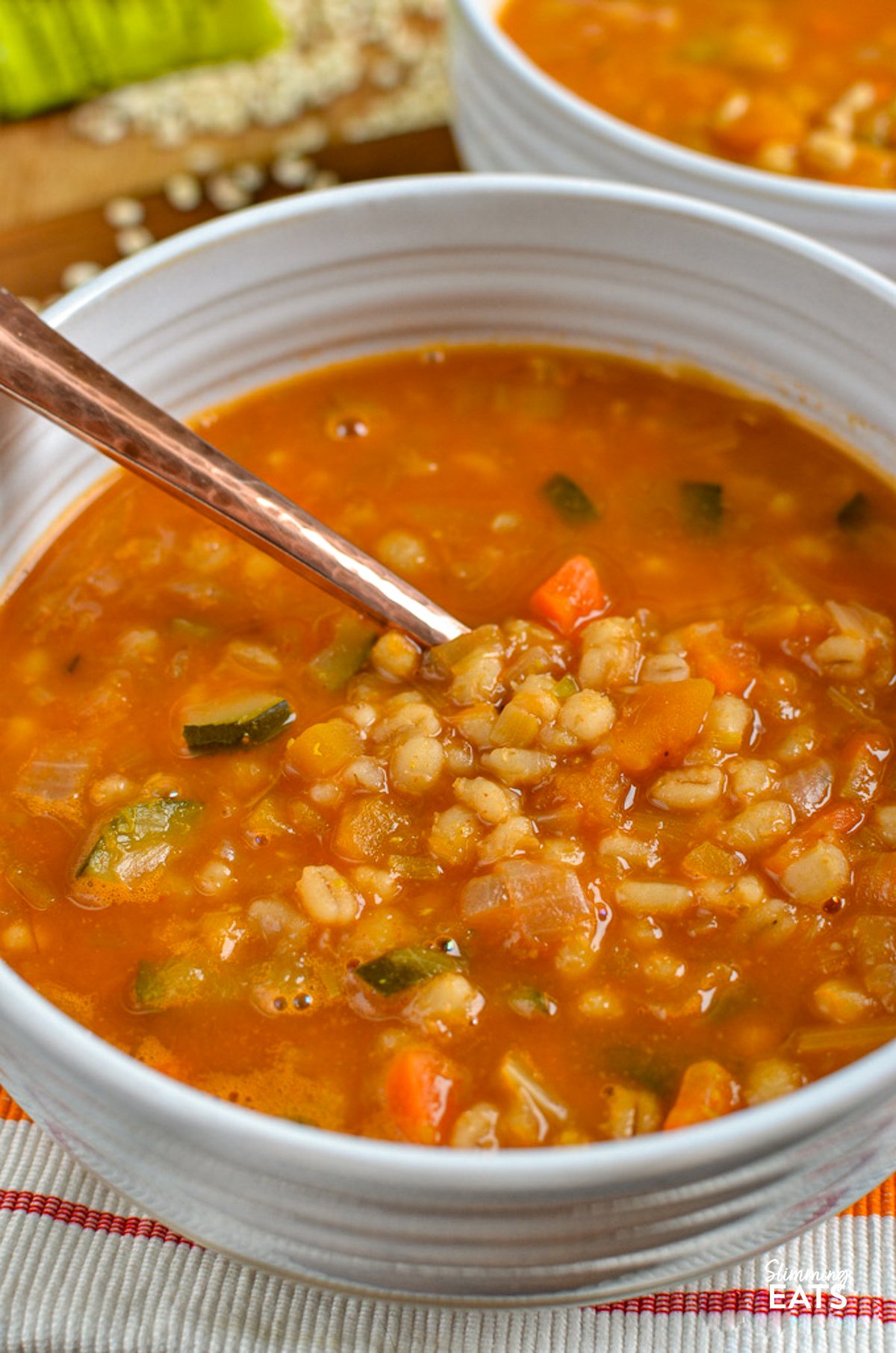 Enjoy the comfort and simplicity of this Vegetable and Pearl Barley Soup - a delightful medley of vegetables teeming with fresh flavours and the wholesome goodness of pearl barley. Vegetable Pearl Barley Soup served in a white bowl accompanied by a silver spoon, with an additional bowl of soup in the background and scattered dried pearl barley grains on the surface around the bowls