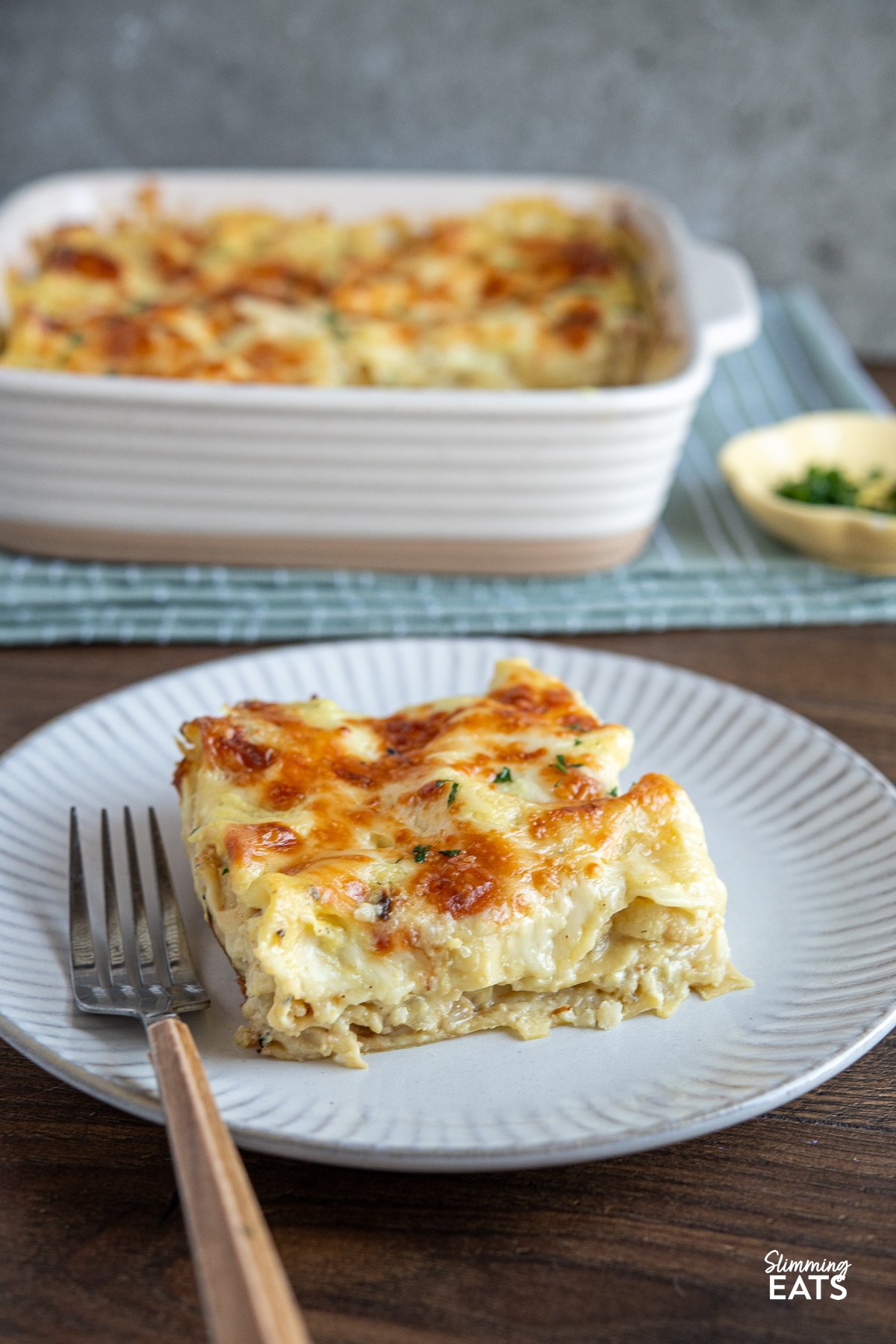 Indulge in the warm, comforting flavours of our Roasted Cauliflower Cheese Lasagne. The sumptuous layers of creamy, rich cheese and the nutty, caramelised cauliflower come together in a harmonious blend that feels like a match made in heaven. Roasted cauliflower lasagne plated on a white grey plate with a fork with wooden-effect handle placed to the left, with the lasagne dish in the background.