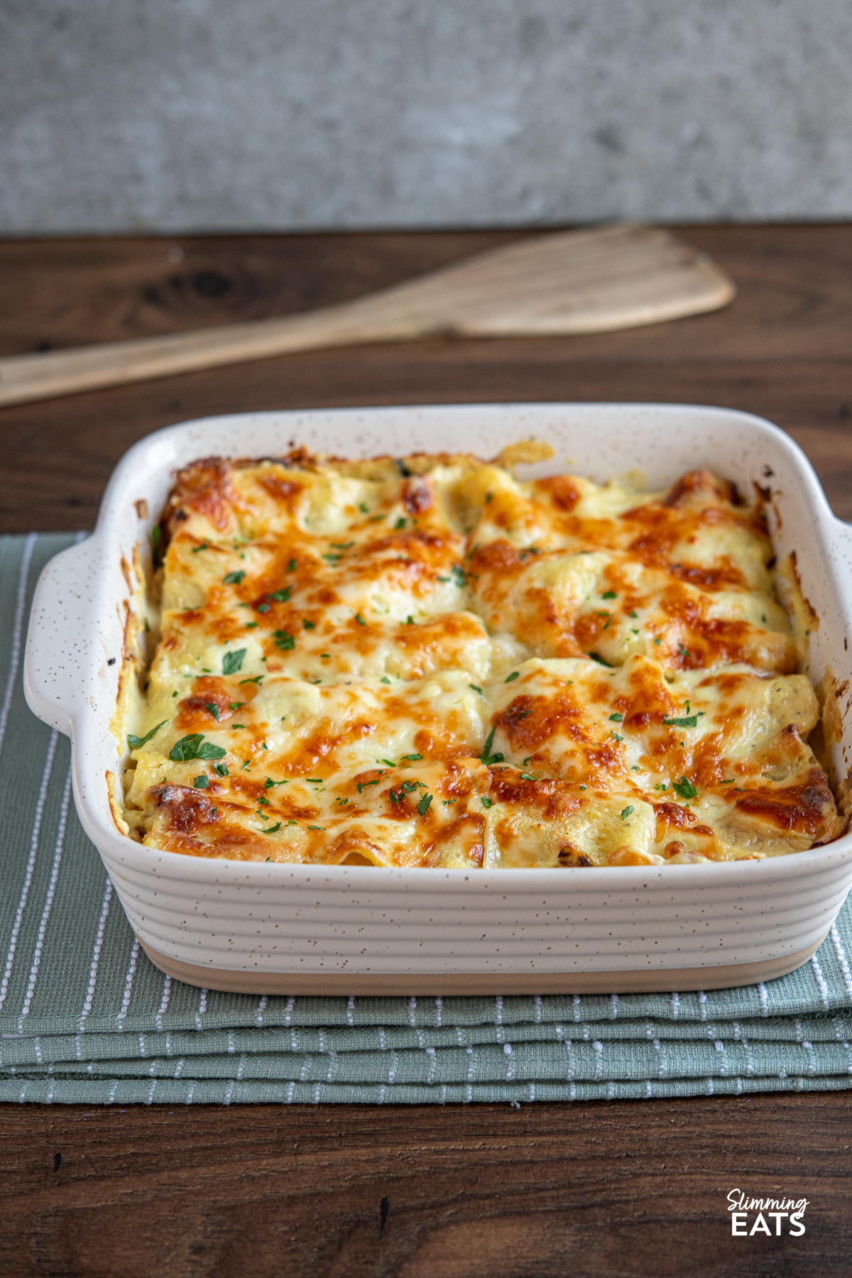 Roasted cauliflower lasagne in a white baking dish  place on a green/gray kitchen towel with a wooden spatula placed behind.