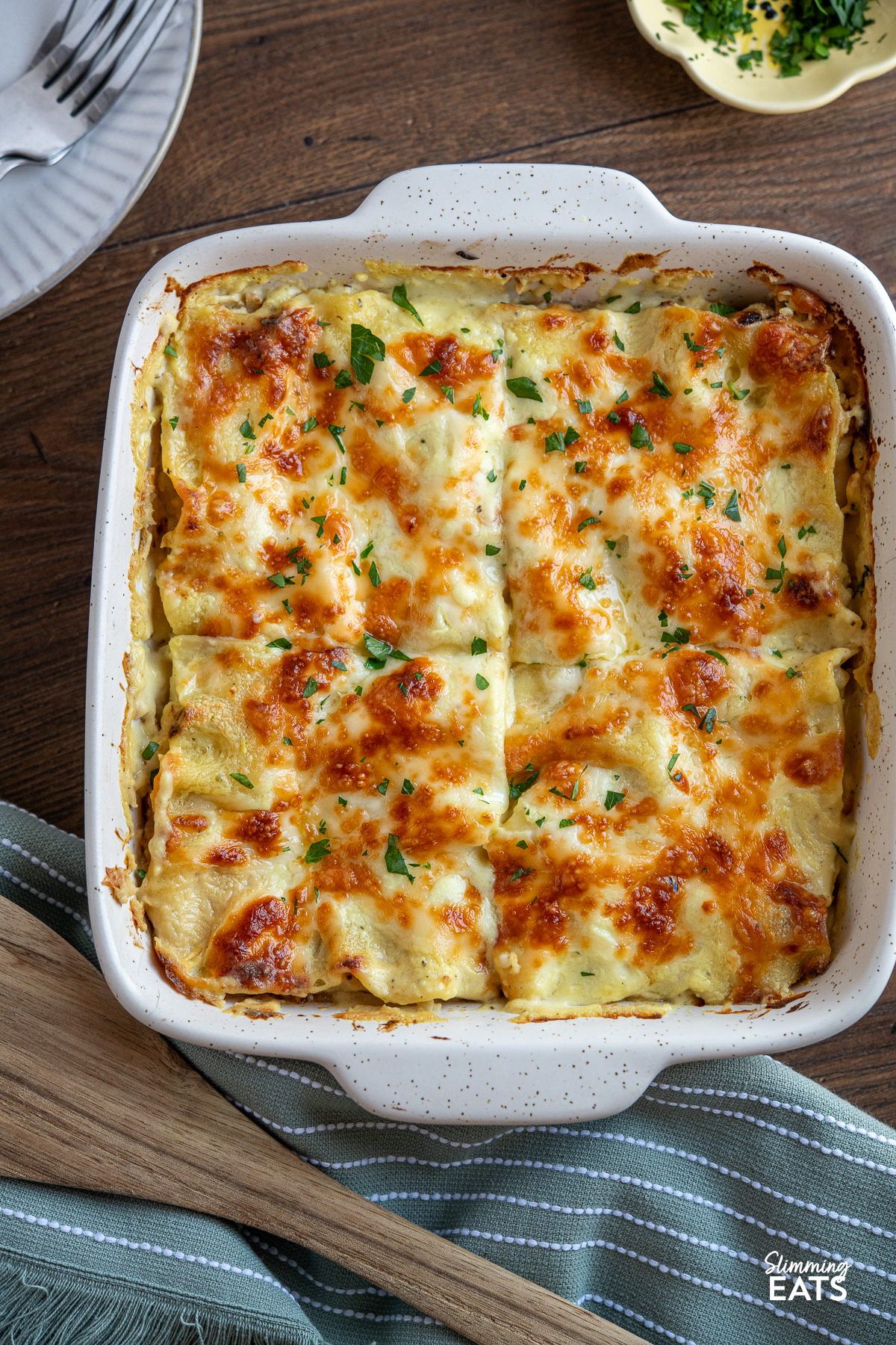 Roasted cauliflower lasagne in a white baking dish with a wooden spatula to the side, accompanied by a plate and a small bowl of herbs in the background, with a green/gray kitchen towel in front.
