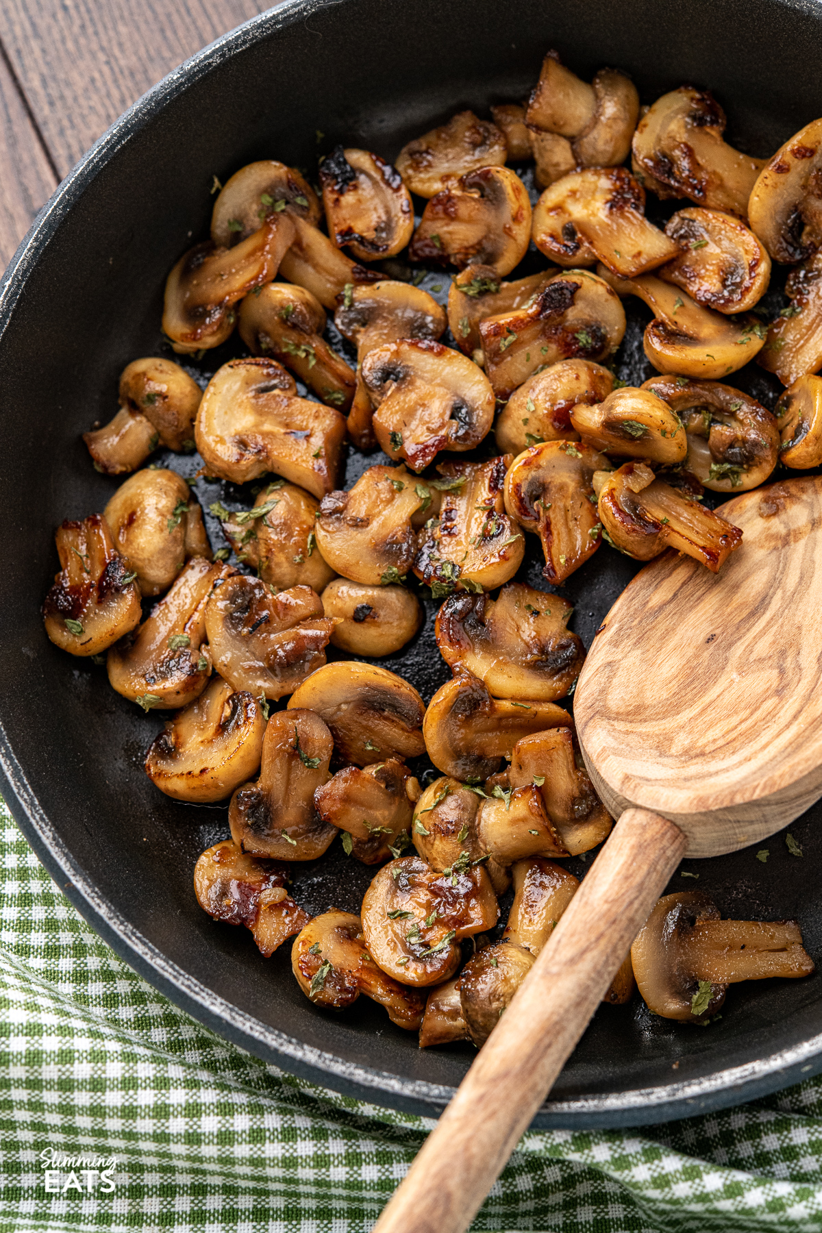 close up of sauteed mushrooms in black frying pan with wooden handle, olive wood spoon placed in pan.