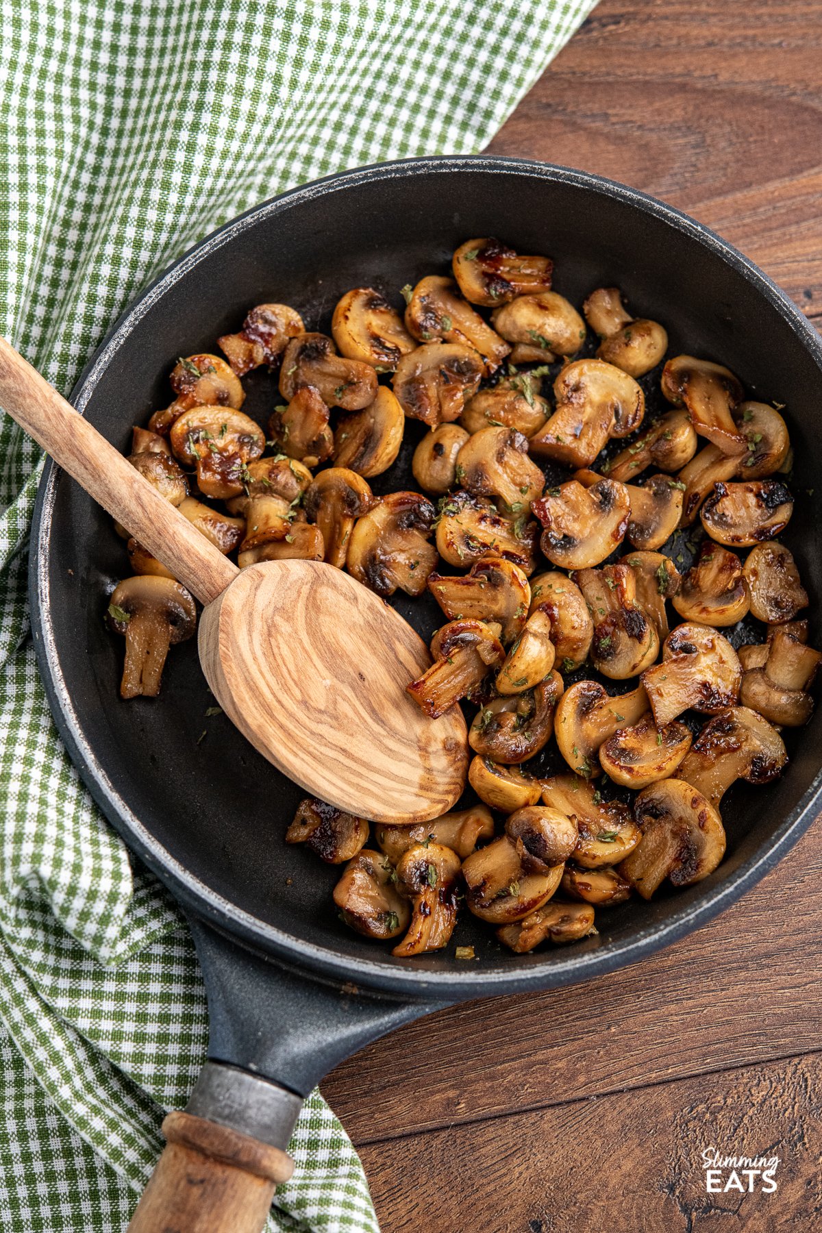 sauteed mushrooms in black frying pan with wooden handle, olive wood spoon placed in pan and green chequed cloth to side. 