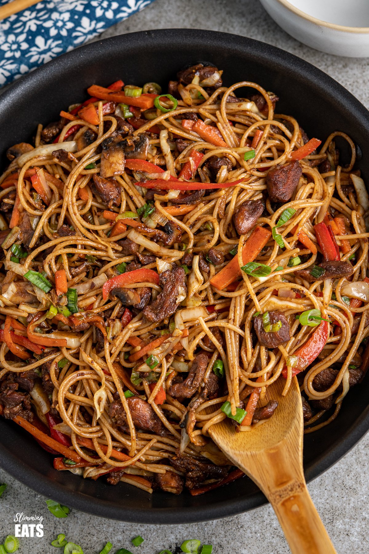 chicken with noodles and vegetables in a black frying pan with scattered spring onions