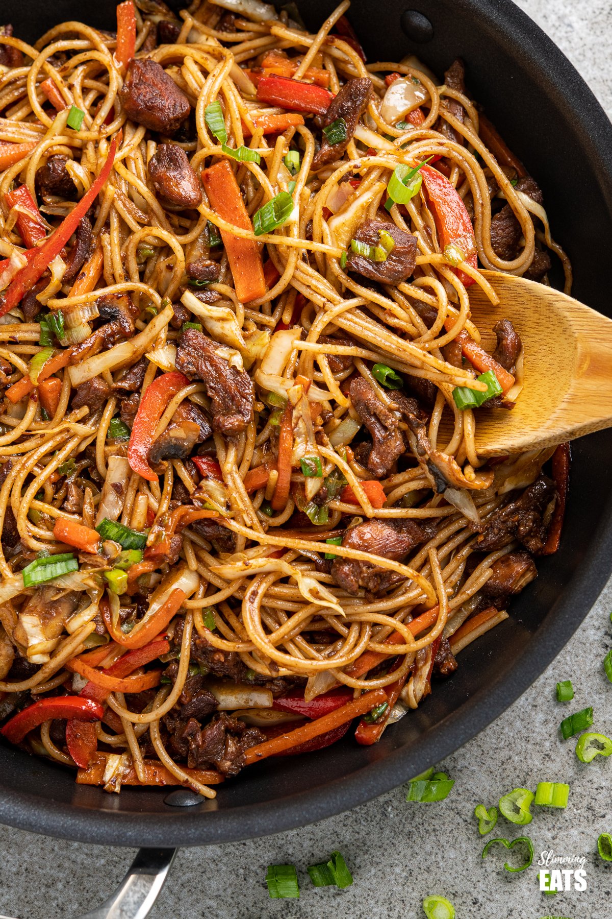 close up of chicken with noodles and vegetables in a black frying pan with scattered spring onions