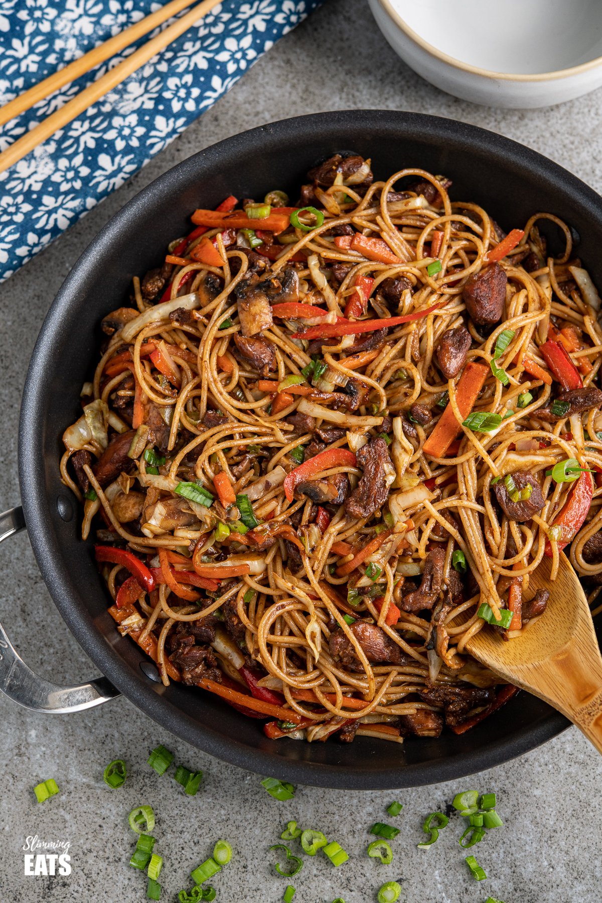 chicken with noodles and vegetables in a black frying pan with scattered spring onions