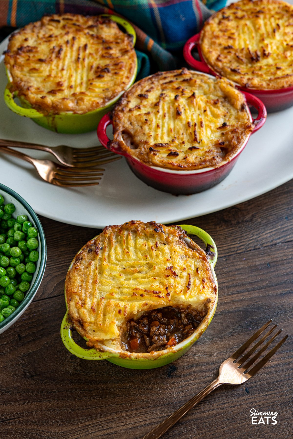 Mini Cottage Pies in a green double handle mini casserole dish surrounded by 3 more mini cottage pies and peas.