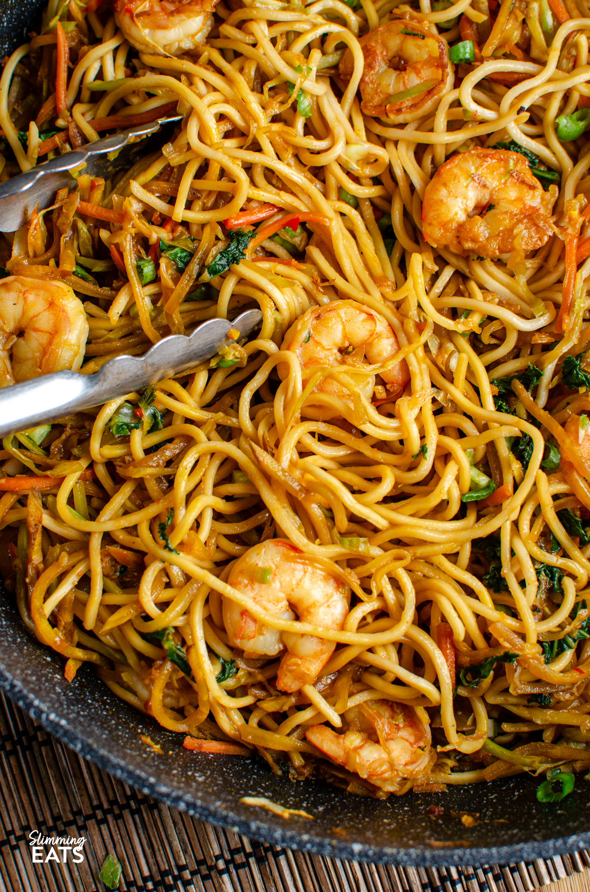 Sweet Chilli Prawn Noodles - an easy dinner that is packed with flavour and ready in less than 30 minutes. Juicy prawns, crisp vegetables and noodles are tossed together in a sweet, chilli sauce with just the right amount of heat for a quick meal that the whole family can enjoy. close up of Sweet Chilli Prawn Noodles in a non stick frying pan with metal tongs