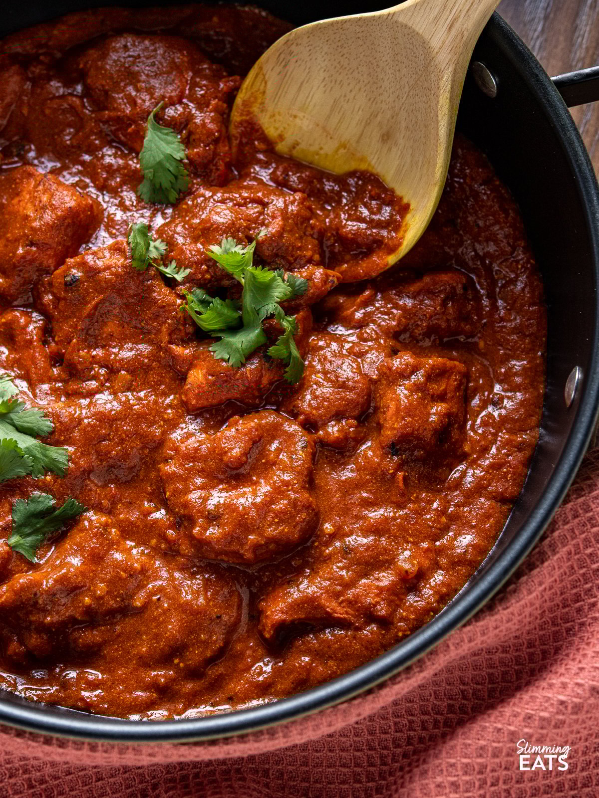 Say goodbye to high calorie takeout and make this delicious low calorie Chicken Tikka Masala. This homemade version will satisfy your cravings without compromising on taste. close up of Lighter Chicken Tikka Masala in black pan with wooden spoon