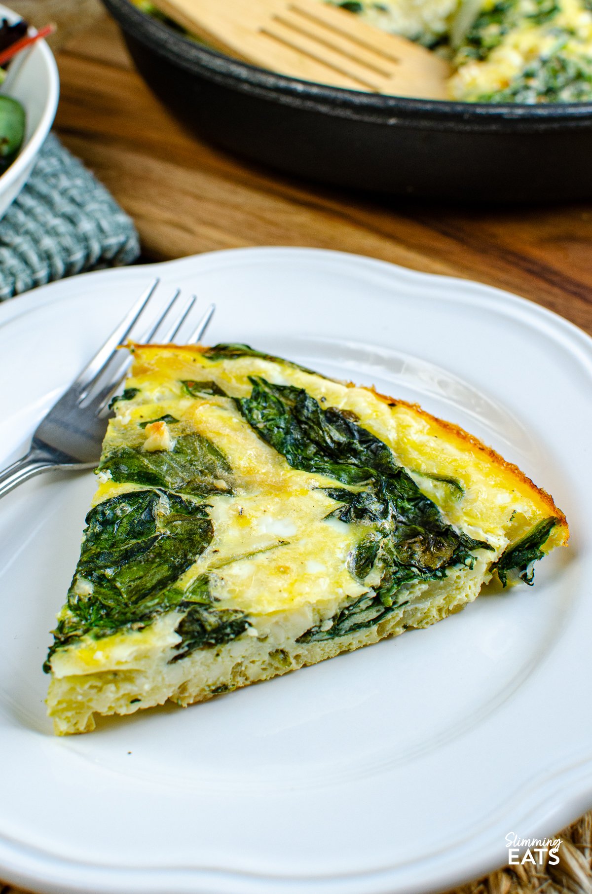 slice of Spinach and Feta Frittata on a white plate, accompanied by skillet with remaining slices in background