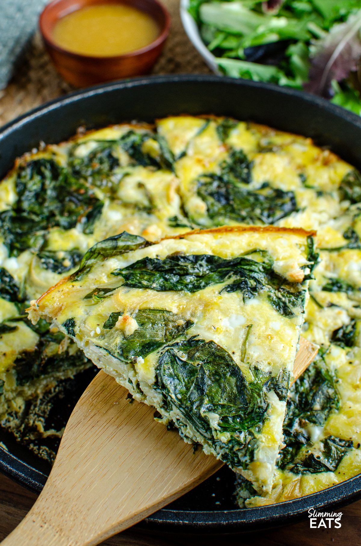 slice of Spinach and Feta Frittata showcased in a skillet, accompanied by a bowl of fresh salad and vinaigrette in the background