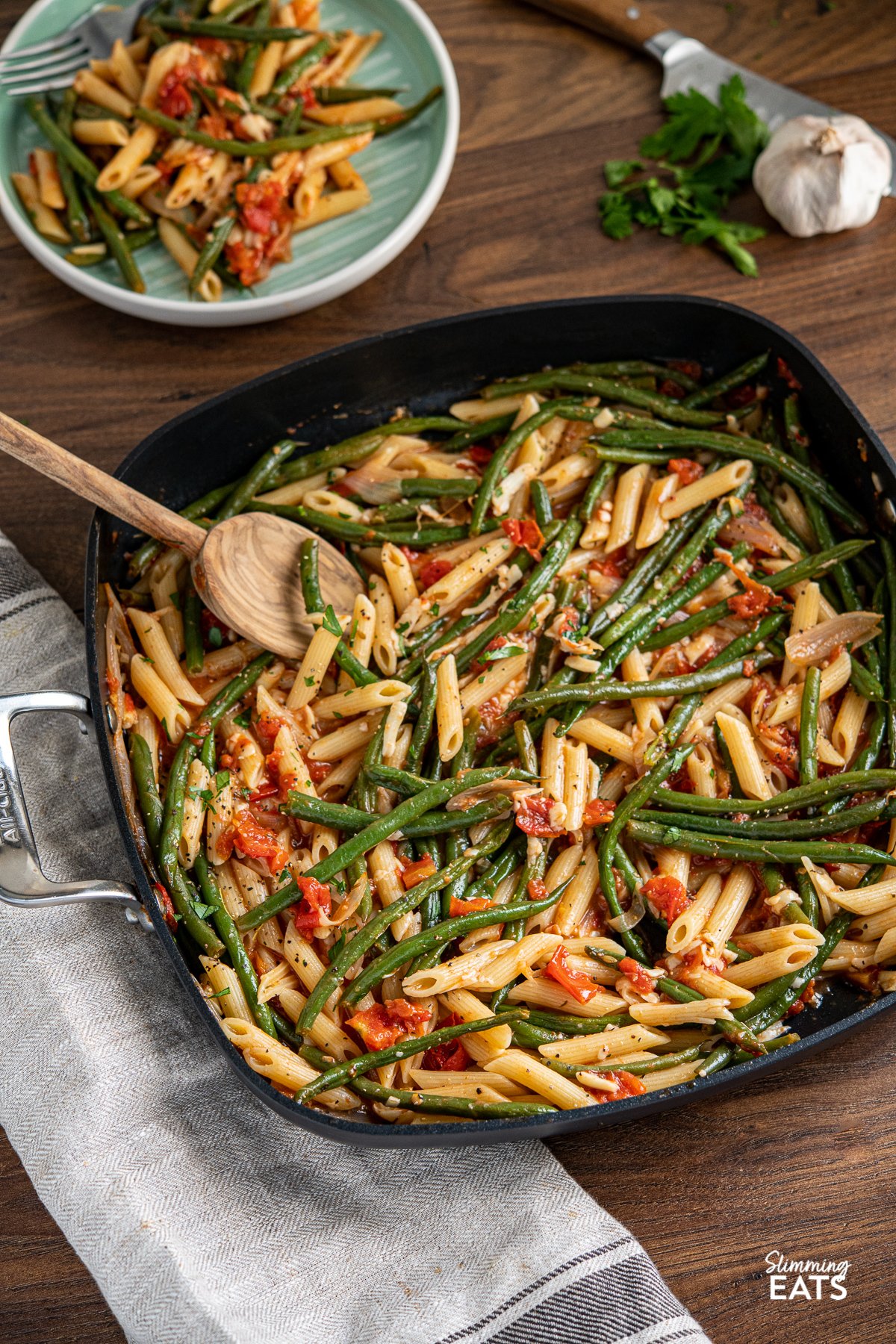 Roasted Green Bean and Tomato Pasta in all clad square skillet with olive wood spoon, fresh garlic and parsley in background as well as plat e with serving of pasta