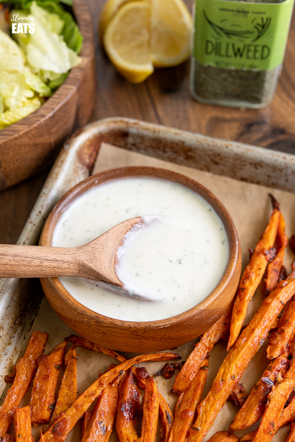 close up of ranch dressing in small wooden bowl next to sweet potato fries on baking tray