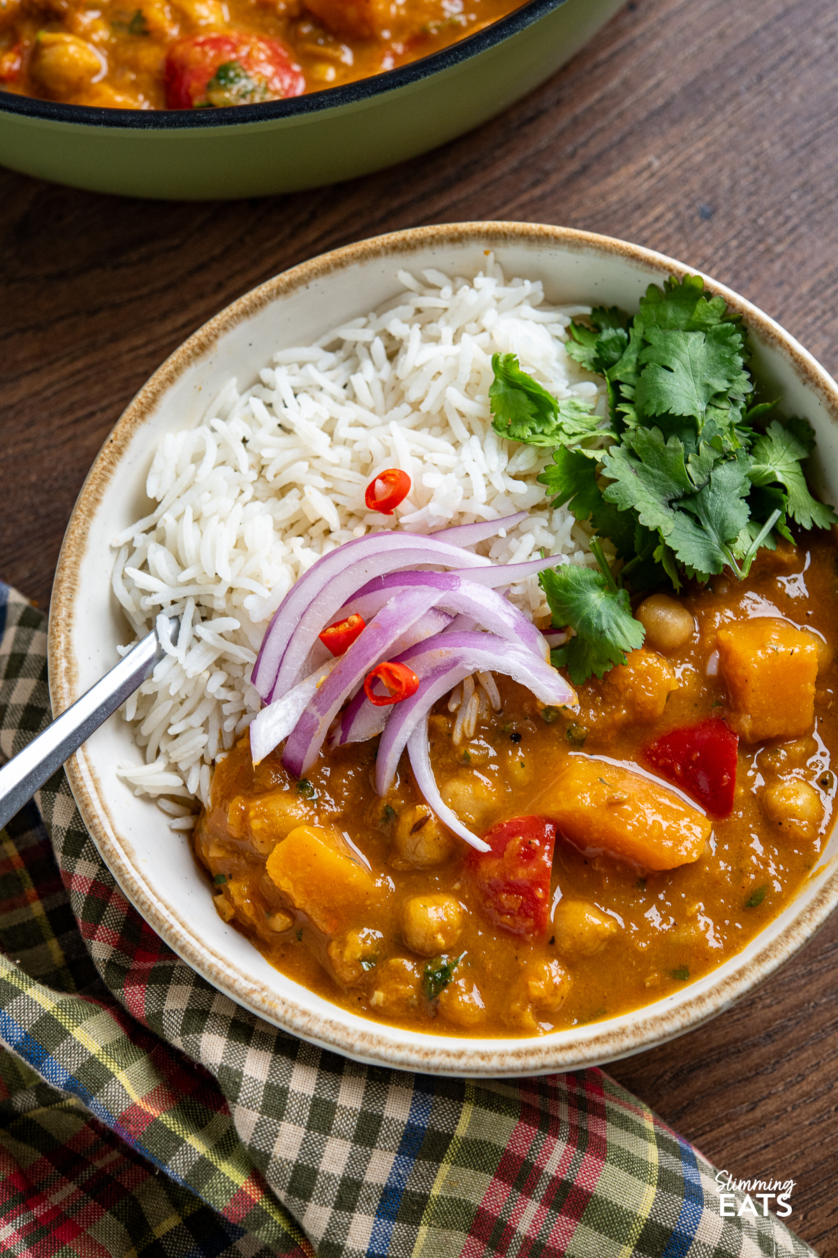 close up of Butternut squash chickpea curry in a small white bowl with a beige rim, accompanied by rice, topped with slices of red chili, red onion, and coriander.