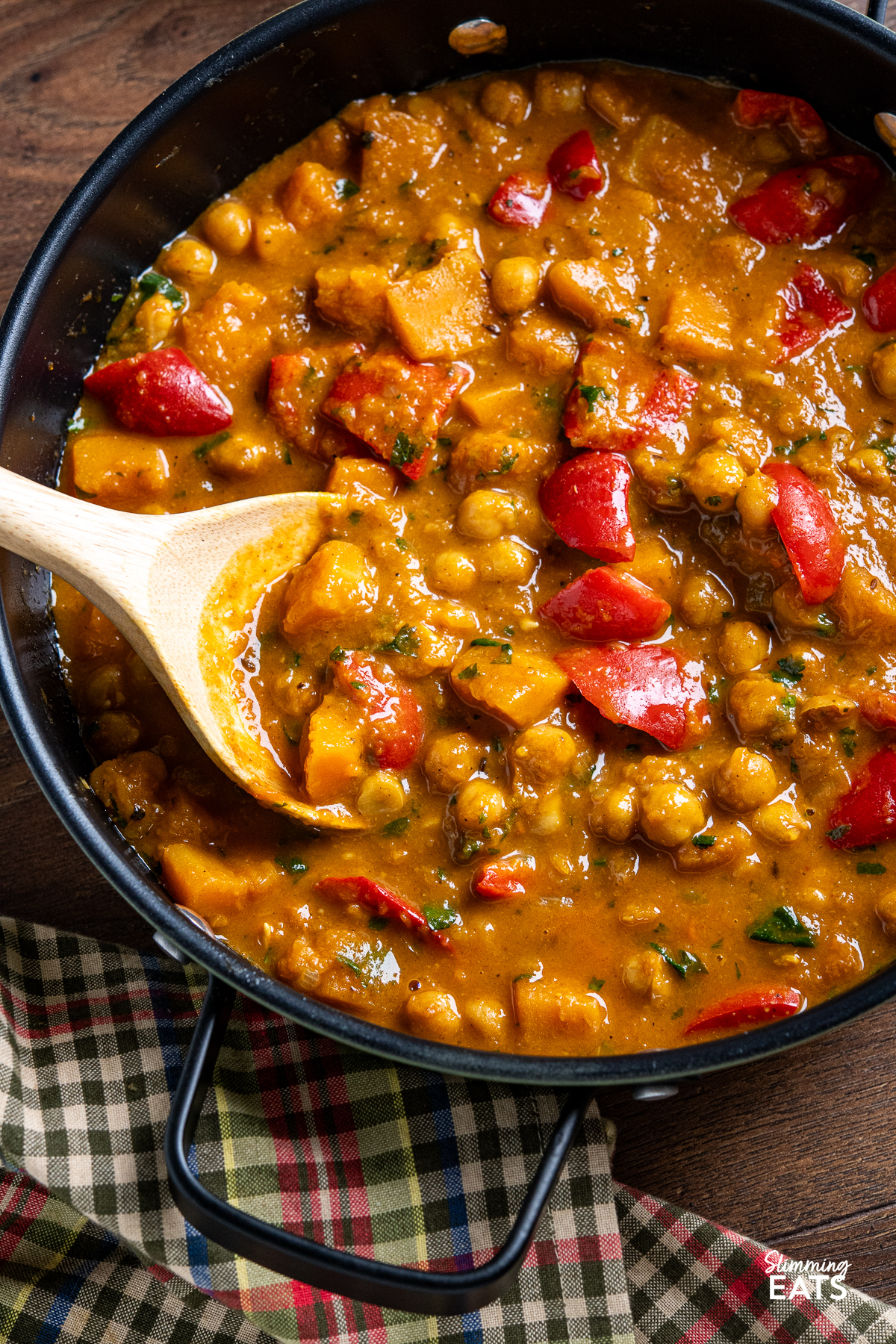 A vibrant chickpea butternut squash curry in a green double-handled frying pan with a wooden spoon