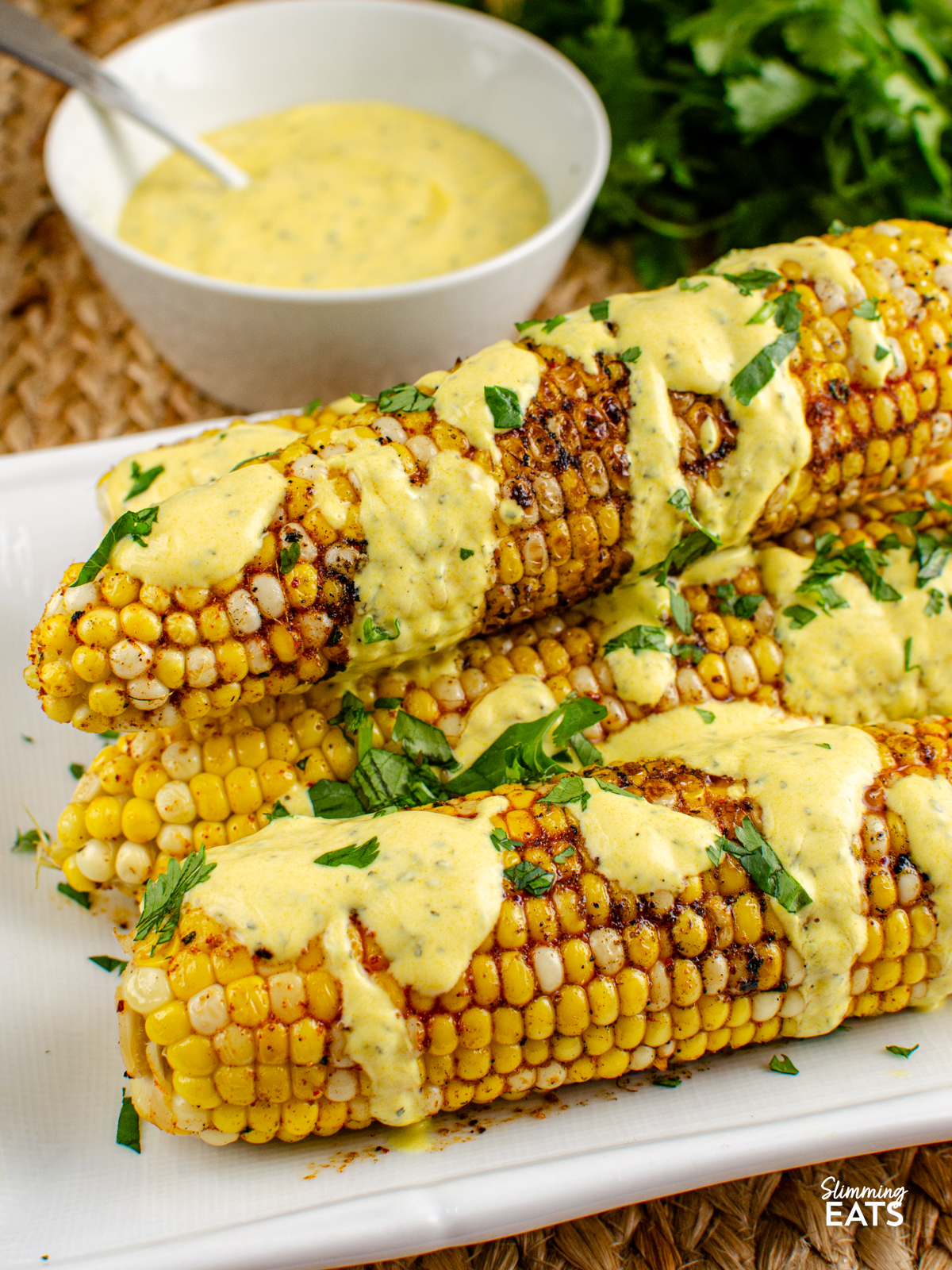 Indian Spiced Corn on the Cob on a white plate drizzled with mint yoghurt, bowl of the yoghurt and fresh coriander next to plate.