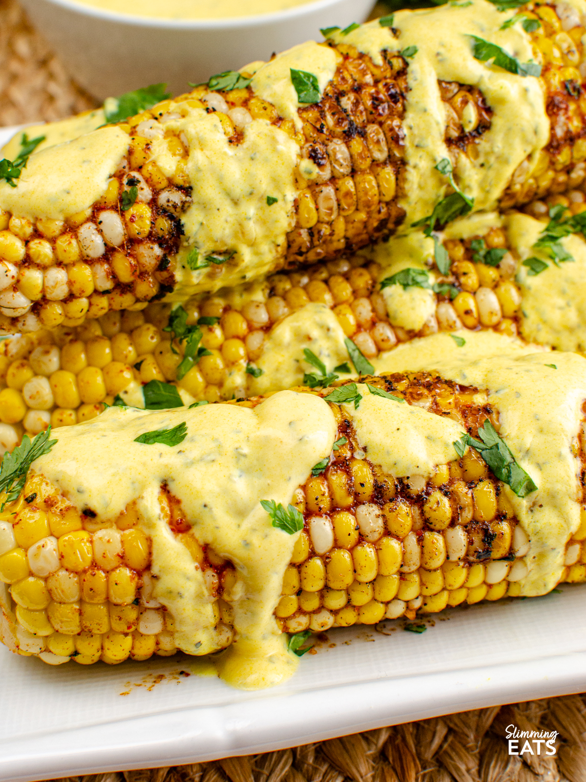Indian Spiced Corn on the Cob on a white plate drizzled with mint yoghurt, bowl of the yoghurt and fresh coriander next to plate.