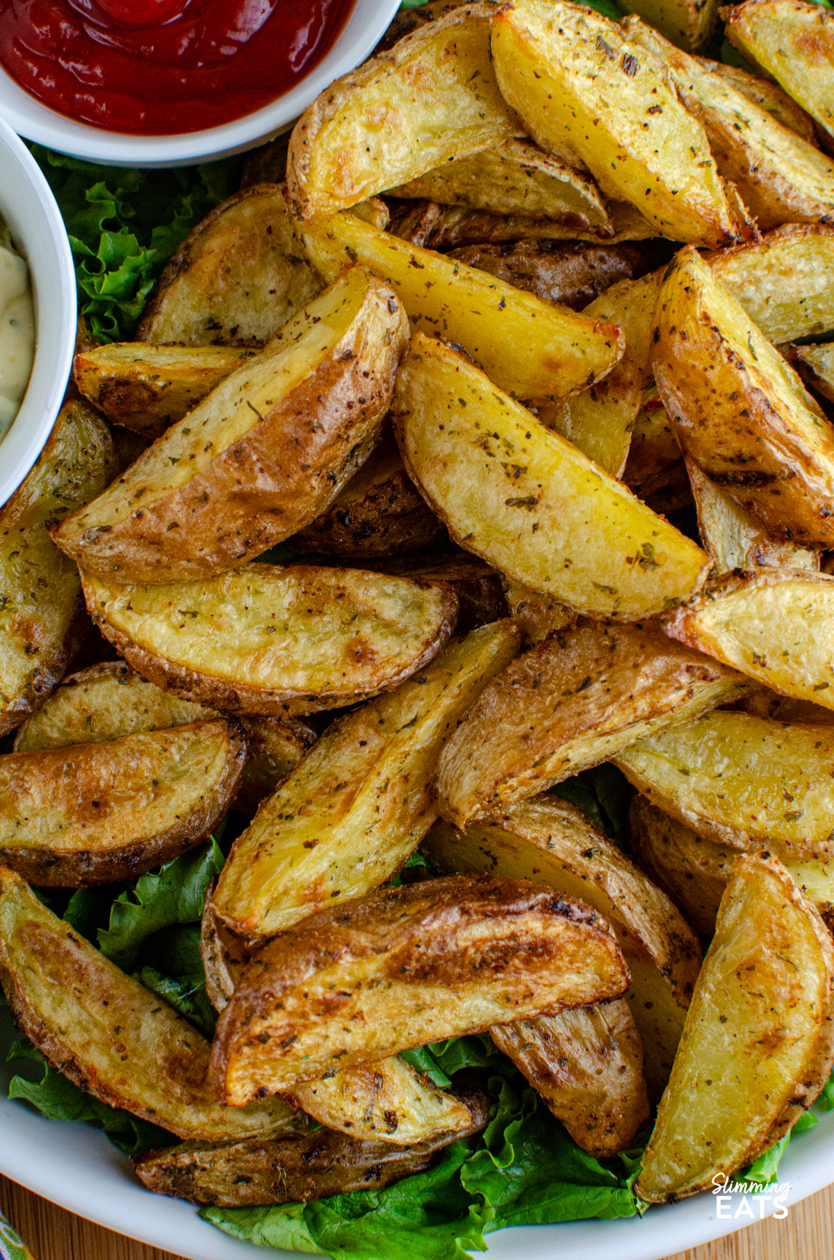 Tuck into these tasty Garlic and Herb Potato Wedges, a perfect side dish to round out just about any meal, and without the guilt too. Crispy on the outside, fluffy in the middle, they’re everything you want from a good wedge and so easy to make at home. close up of Garlic and Herb Potato Wedges in a bowl with lettuce, ketchup and garlic mayo.
