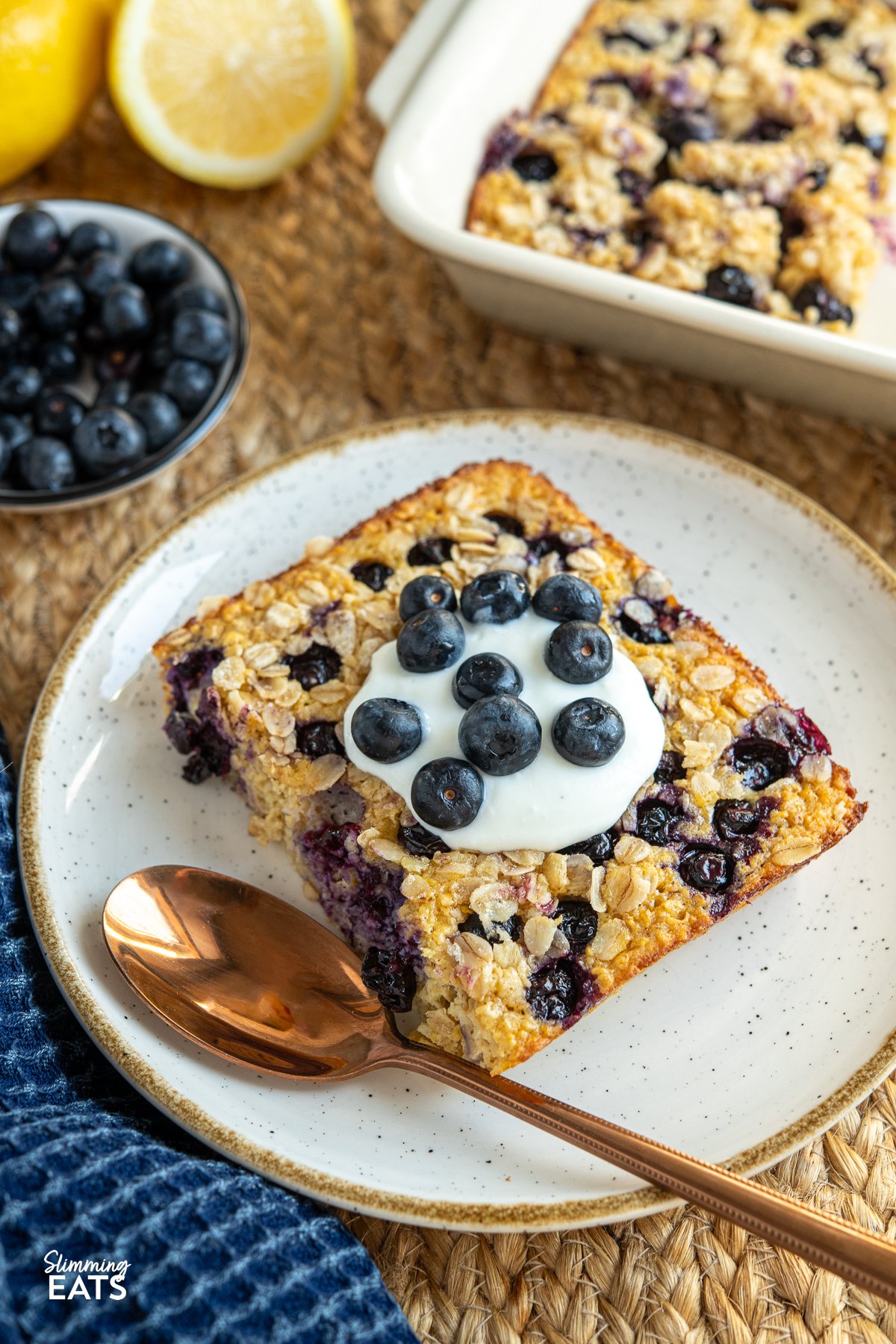 serving of lemon blueberry baked oats topped with yoghurt and blueberries on a white plate with mustard coloured rim, spoon place to the left.