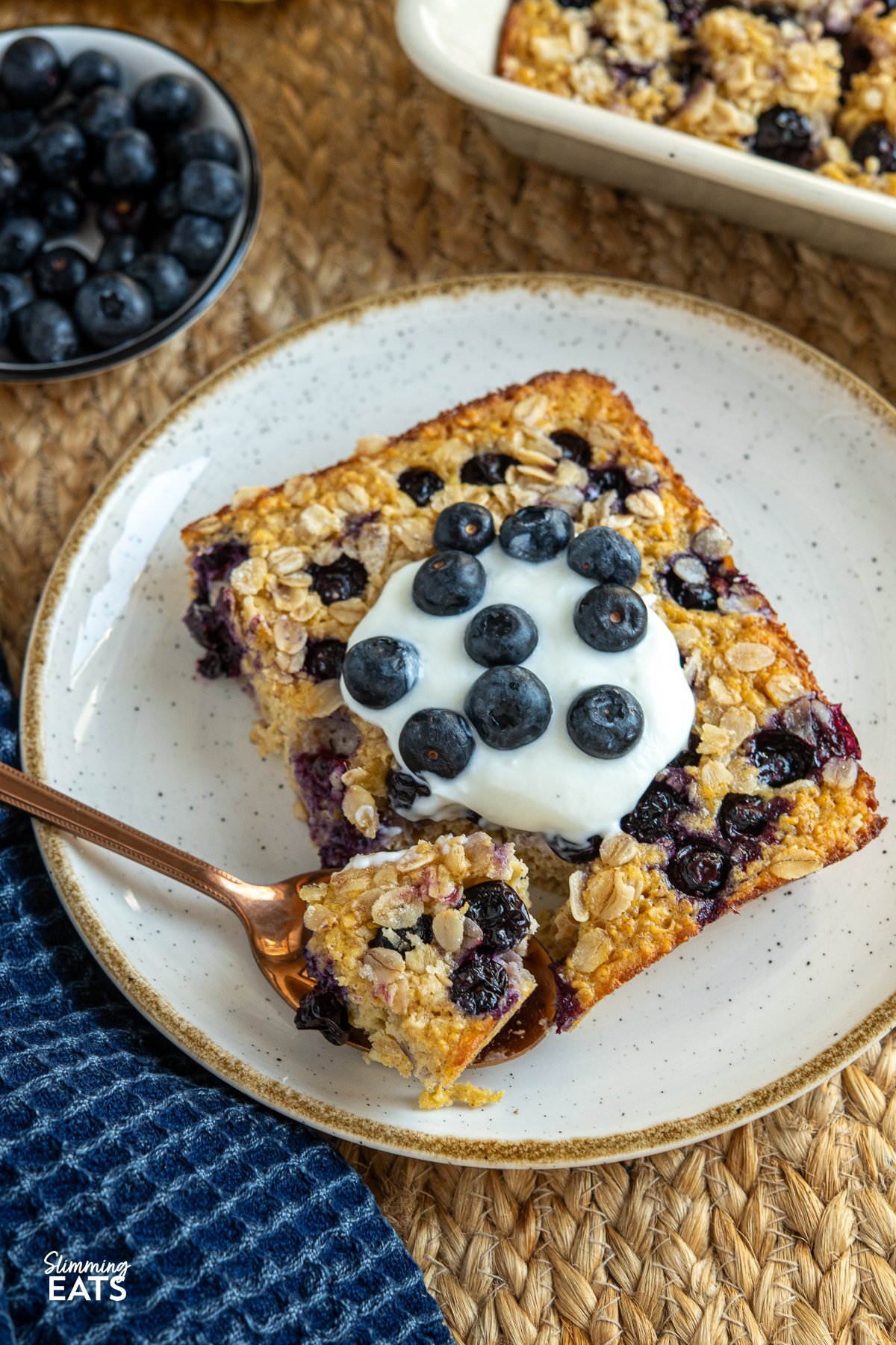 close up of serving of lemon blueberry baked oats topped with yoghurt and blueberries on a white plate with mustard coloured rim, spoon place to the left.