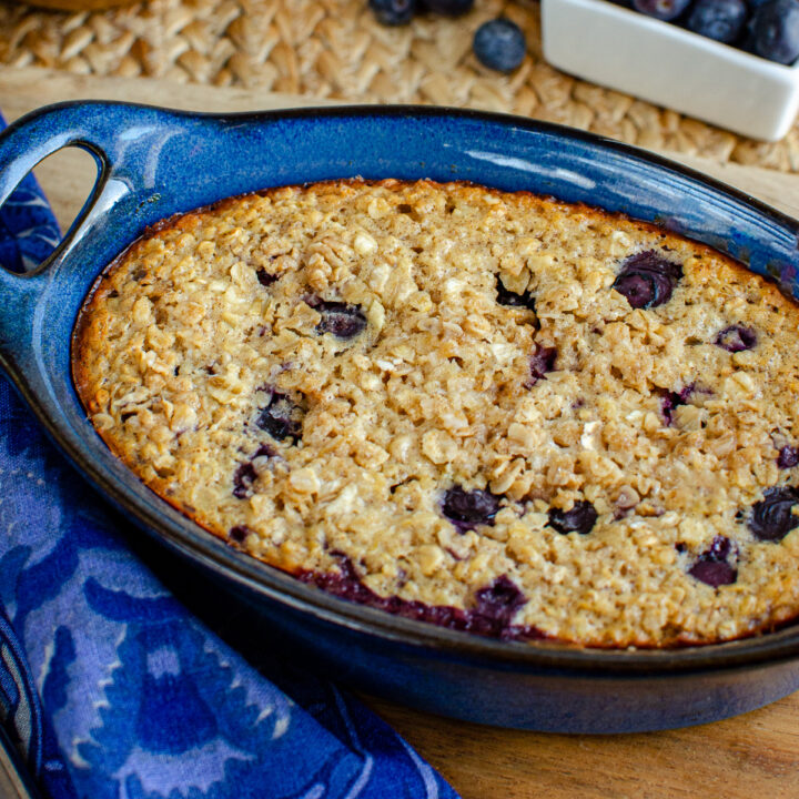 Lemon blueberry baked oats in a blue ceramic dish, showing a golden-brown crust sprinkled with oats and sweetener, with hints of lemon zest and blueberries peeking through, ready to be served.
