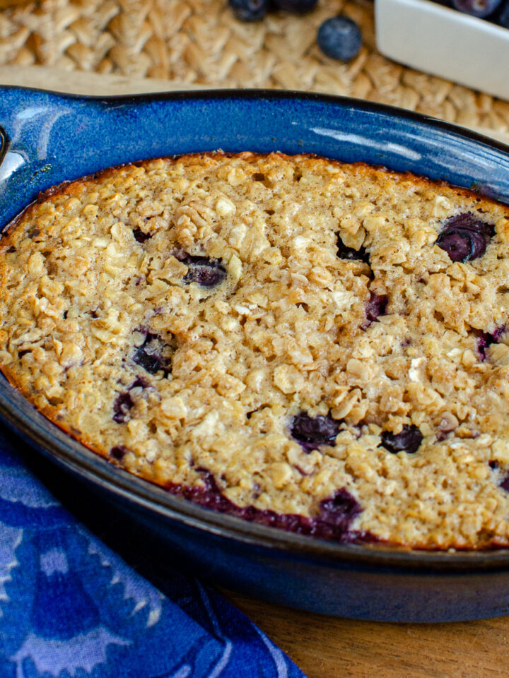 Lemon blueberry baked oats in a blue ceramic dish, showing a golden-brown crust sprinkled with oats and sweetener, with hints of lemon zest and blueberries peeking through, ready to be served.