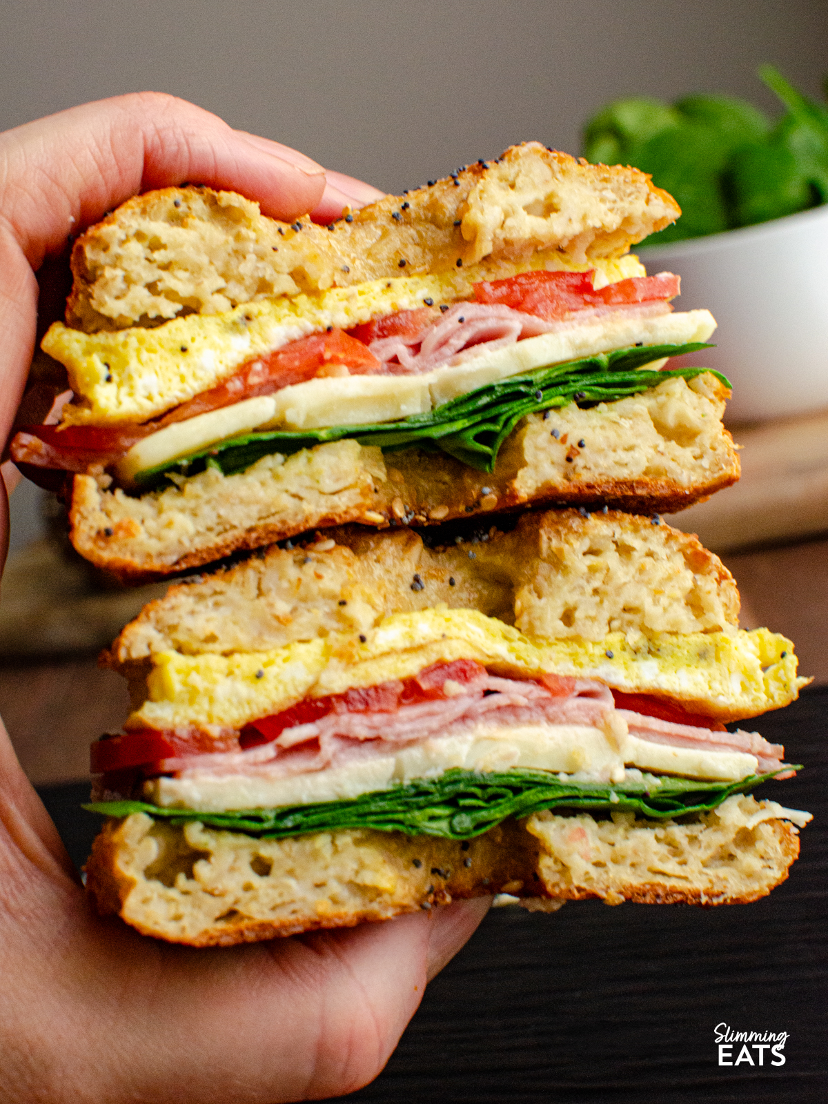 oat bagel sliced in half and filled with egg, cheddar, tomato and baby spinach being picked up by hand ready to eat. 