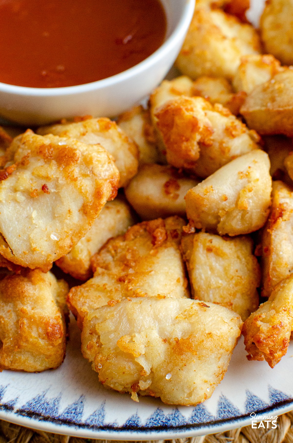 Forget takeout with this delicious Sweet and Sour Chicken Bites that can be cooked in an Air Fryer or Oven and are the perfect addition to your Chinese fakeaway night. close up Sweet and sour chicken bites on a plate with a blue and navy patterned rim, accompanied by a small bowl of sweet and sour sauce on the side.