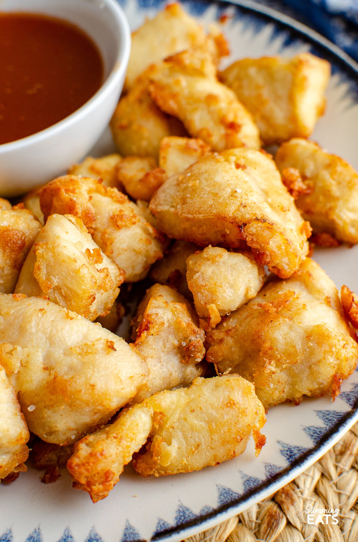 Forget takeout with this delicious Sweet and Sour Chicken Bites that can be cooked in an Air Fryer or Oven and are the perfect addition to your Chinese fakeaway night. close up of Sweet and sour chicken bites on a plate with a blue and navy patterned rim, accompanied by a small bowl of sweet and sour sauce on the side.