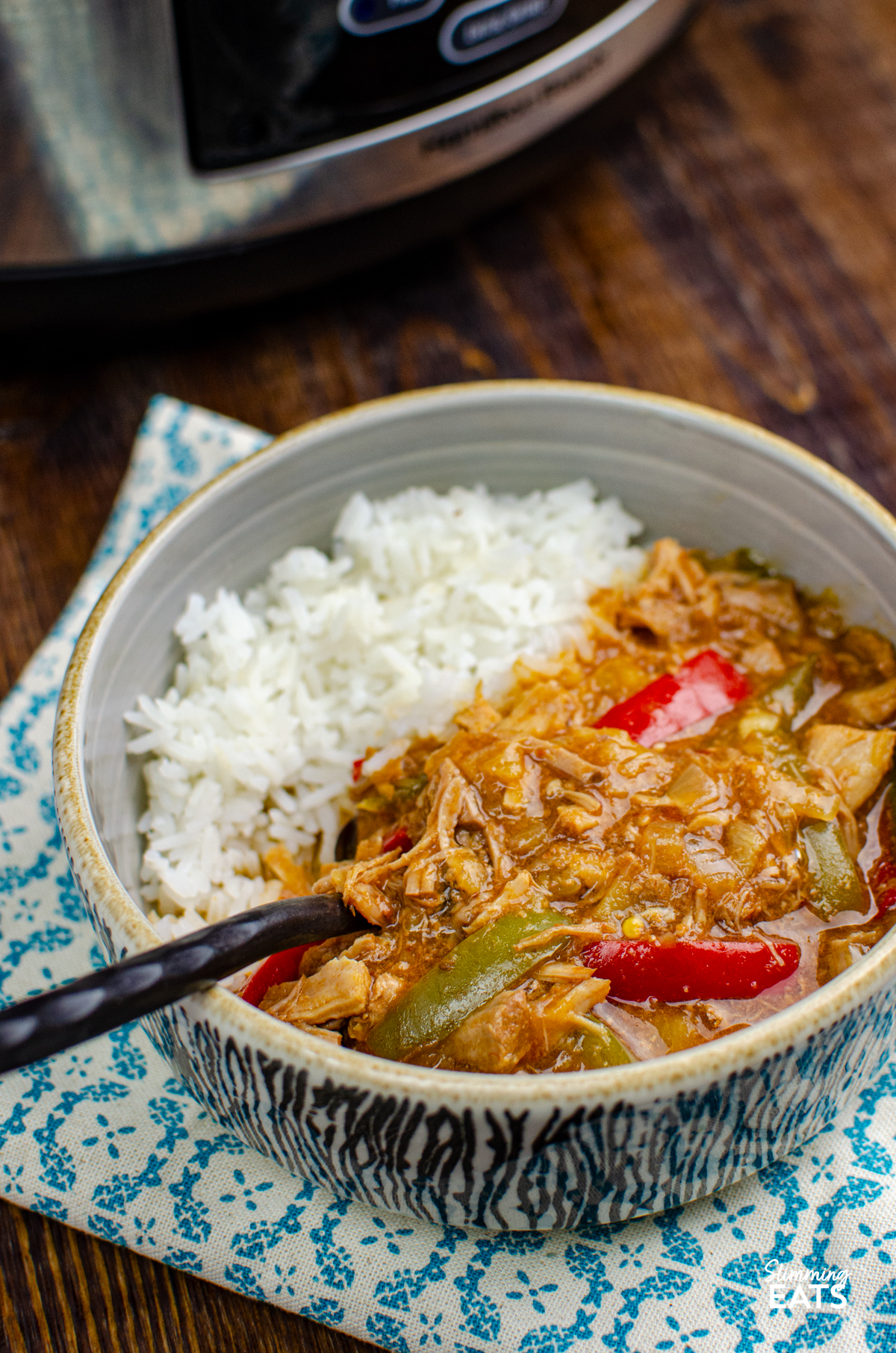 Slow Cooker Pineapple Pork - tender slow cooked pork in a delicious sweet and savoury sauce with pineapple, peppers, soy sauce and a little kick of spice. It is the perfect easy dinner that is ideal for busy days when you want dinner ready and waiting for you when you get home. serving of Slow Cooker Pineapple Pork with rice in grey bowl with black and white pattern, fork placed in bowl and slow cooker visible in background