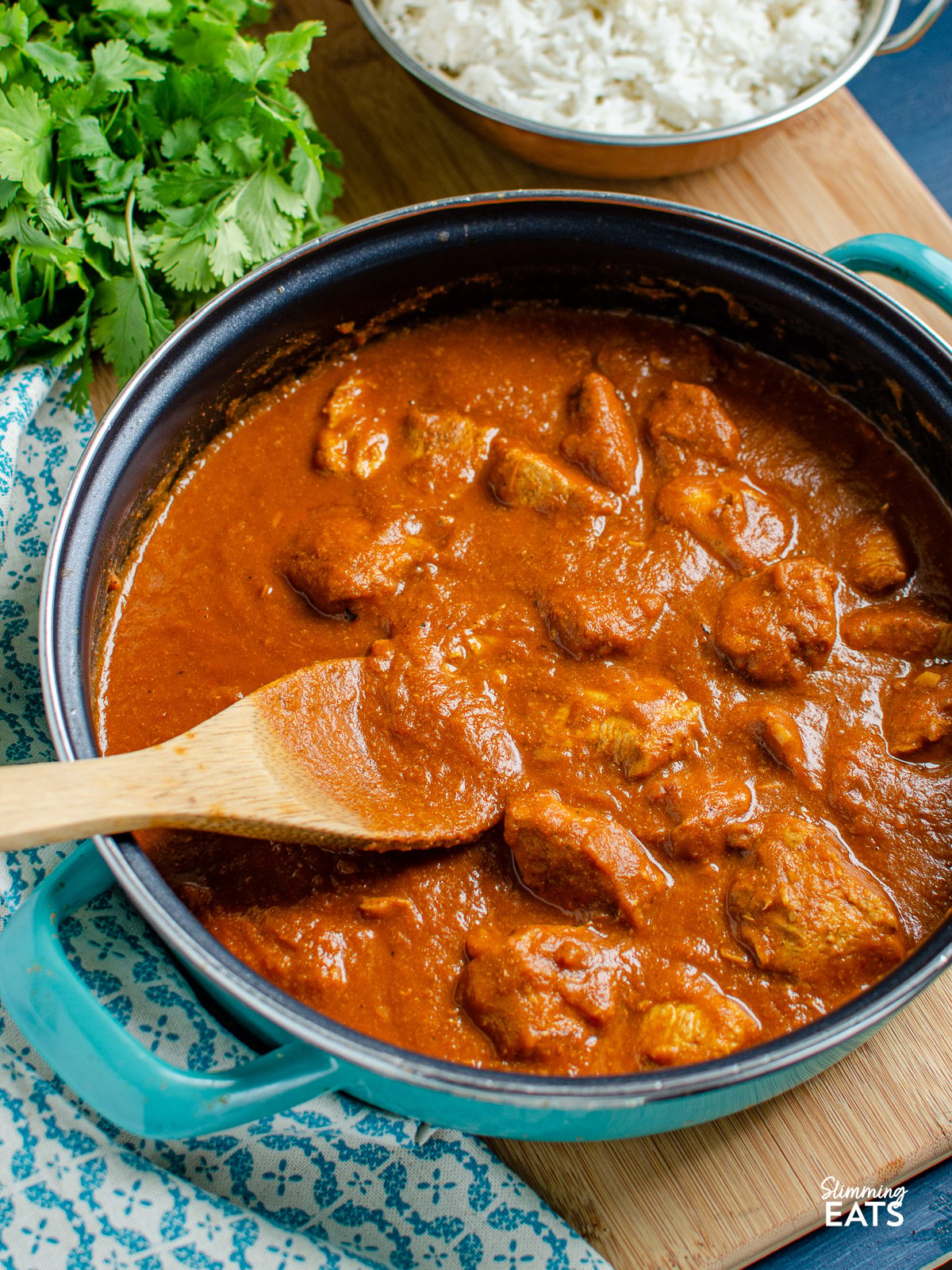 Butter Chicken in a non stick frying pan with wooden spoon and rice and coriander in background