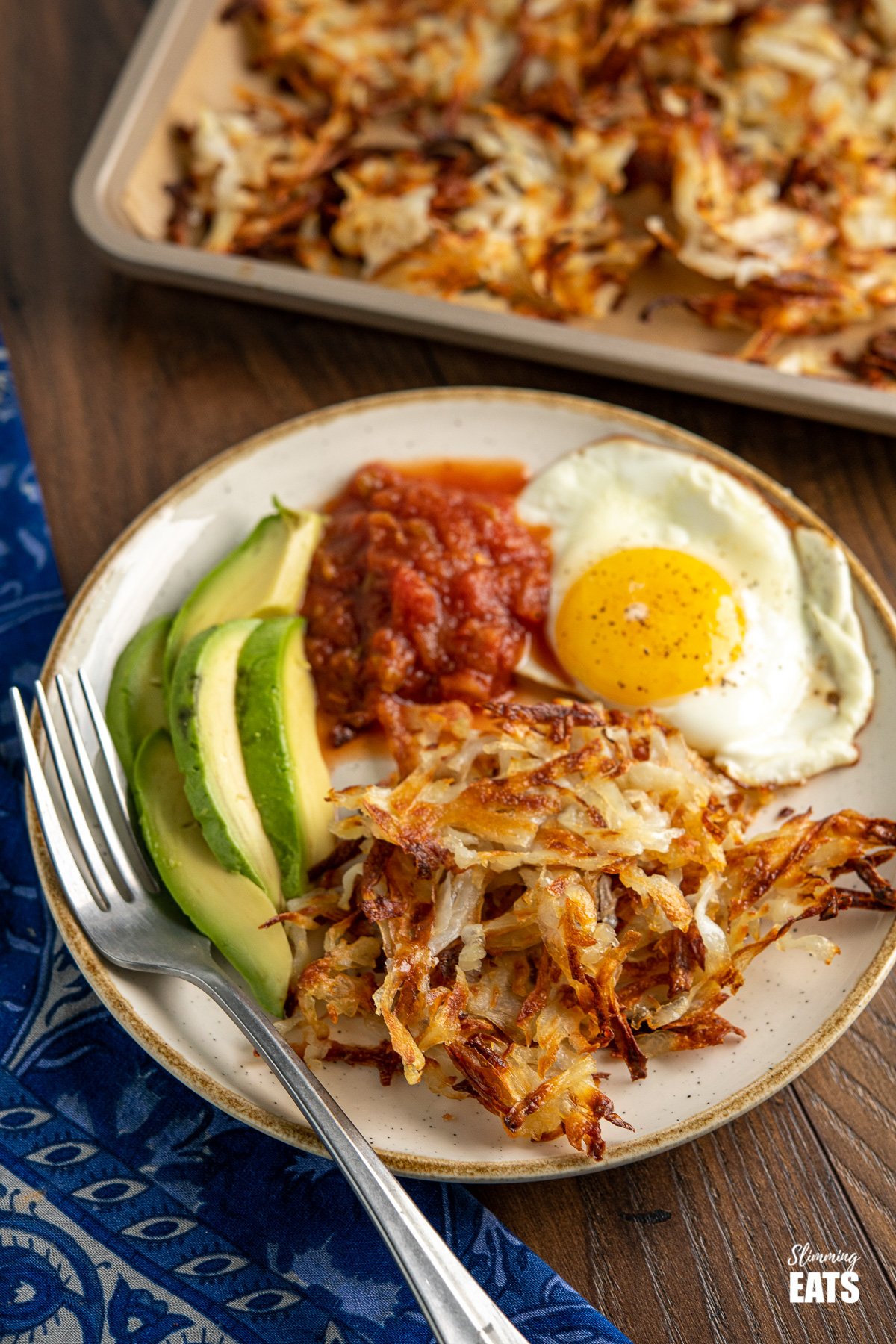 shredded potato hash browns on white plate with egg, salsa and avocado, fork placed to the side and tray of hash browns in the background