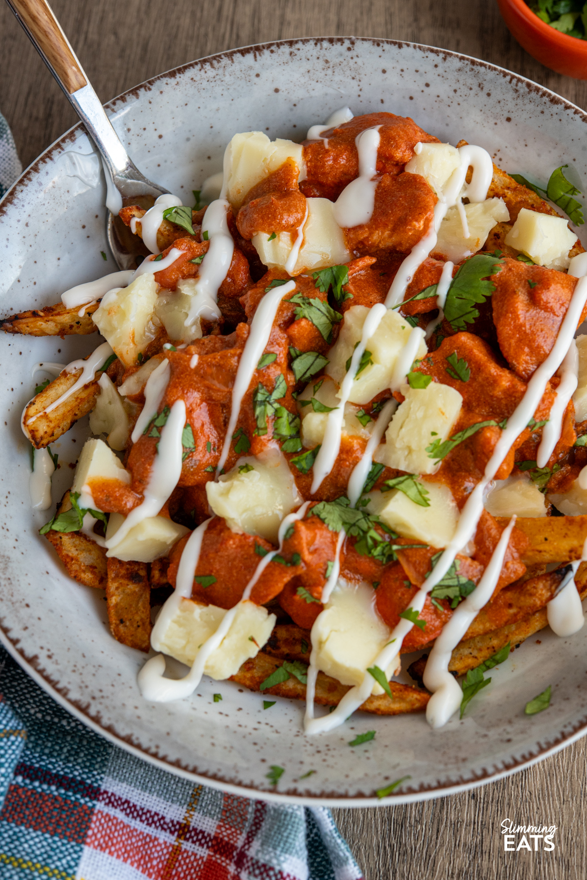 Butter Chicken Poutine!! - The ultimate dish for leftover butter chicken with crispy fries, melty cheese and plenty of flavour. Butter Chicken Poutine in a brown grey bowl with fork, small orange bowl of coriander next to bowl.