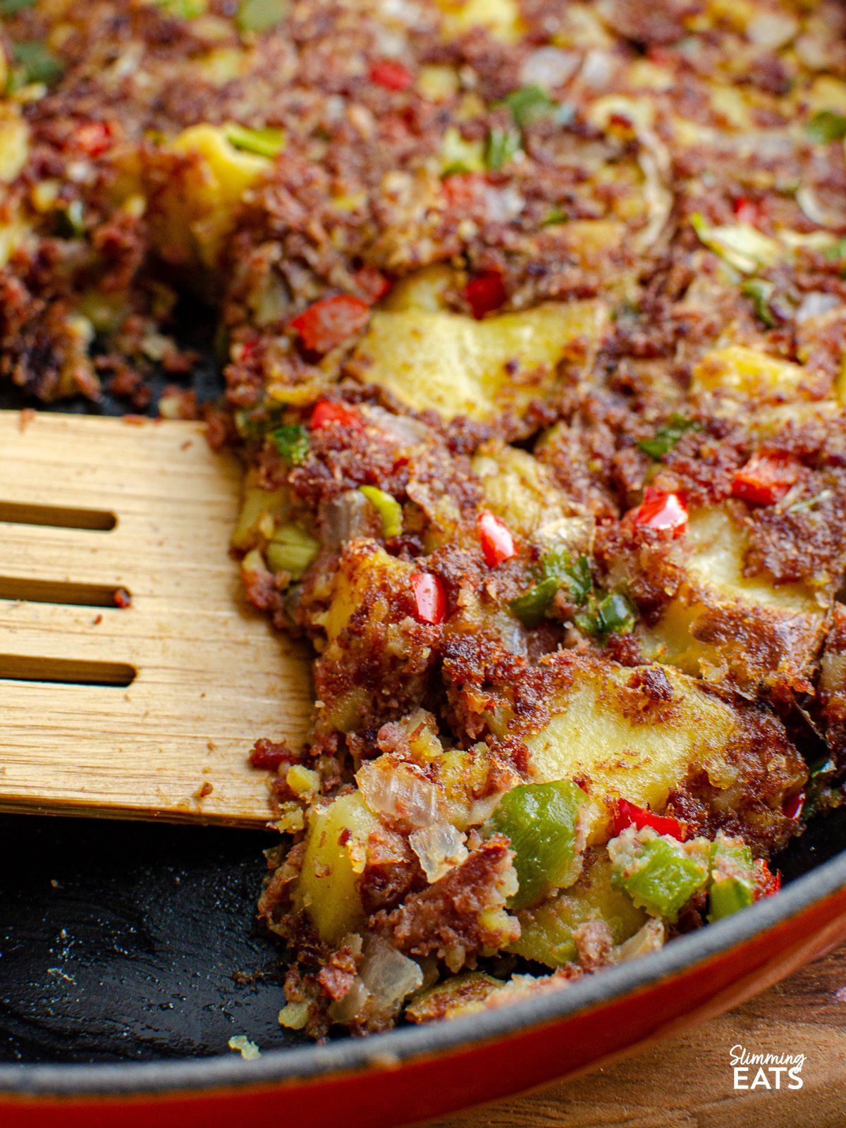 Corned Beef Hash Skillet with peppers, onion and golden potatoes for an easy family friendly recipe that is perfect for breakfast, lunch or dinner wooden spatula scooping Corned Beef Hash in a cast iron orange Le Creuset skillet