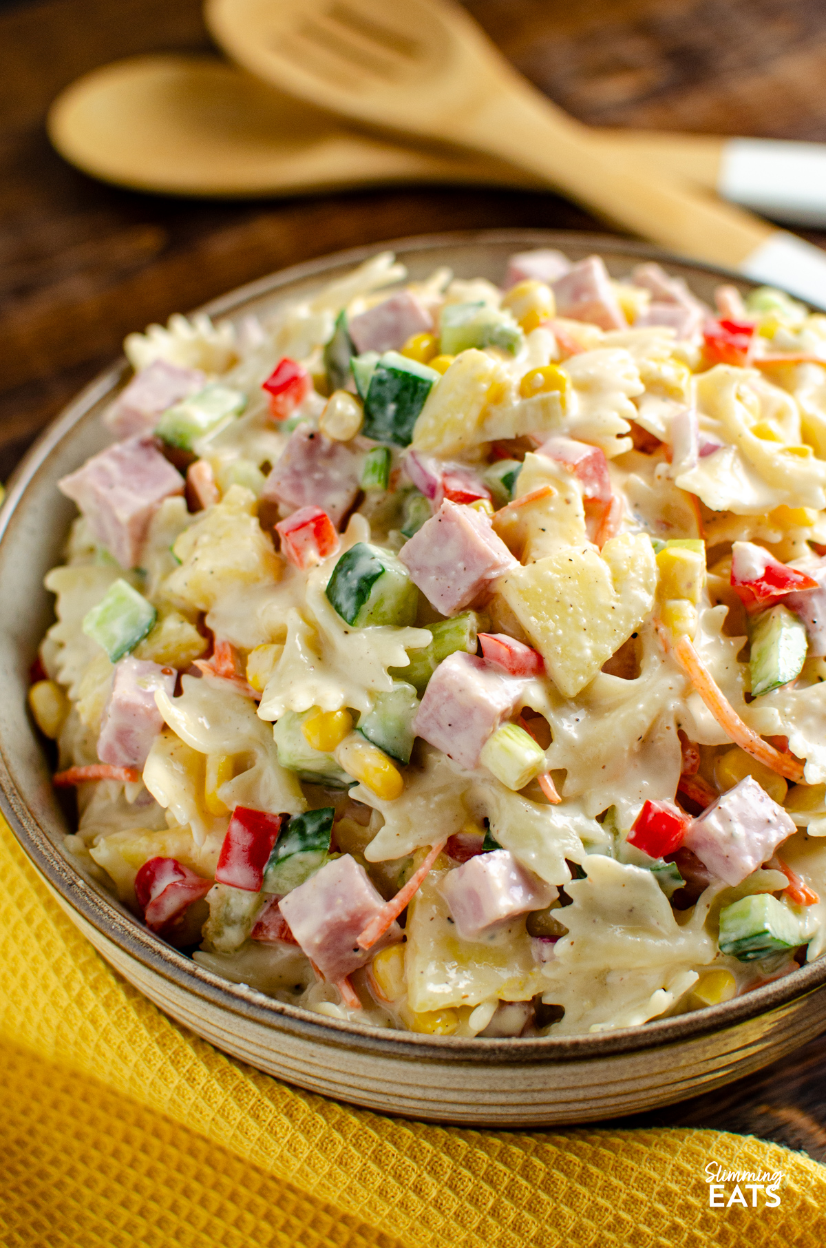This Hawaiian Pasta Salad is a fresh, creamy and colourful side dish packed with ham, pineapple and crunchy vegetables, perfect for barbecues, picnics or easy lunches. hawaiian pasta salad in a big serving bowl with wooden salad spoons in background and yellow placemat in front