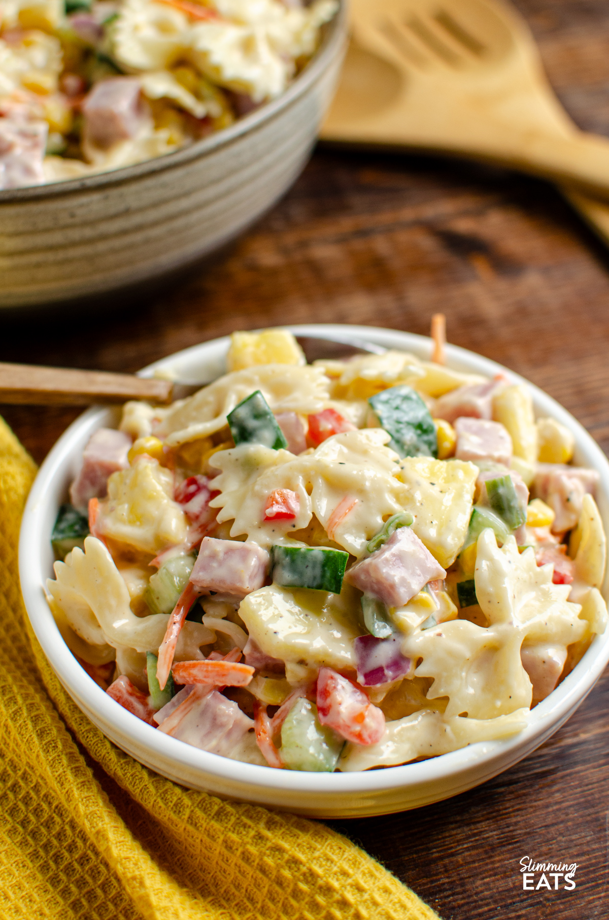This Hawaiian Pasta Salad is a fresh, creamy and colourful side dish packed with ham, pineapple and crunchy vegetables, perfect for barbecues, picnics or easy lunches. serving of Hawaiian Pasta Salad in a white bowl with spoon, big serving bowl with the salad in background with wooden salad spoons