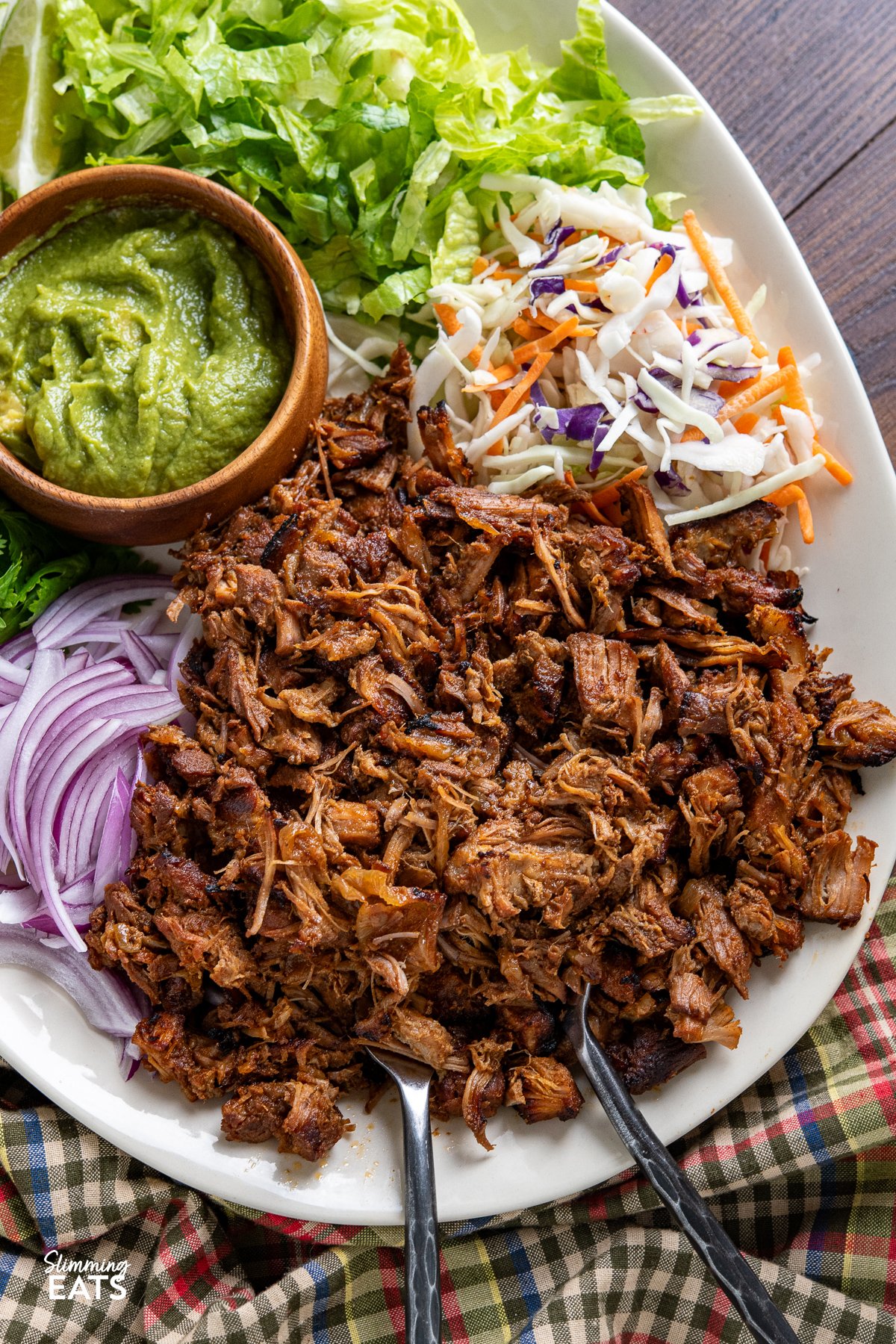 Mexican Pineapple Pork Carnitas on large oval plate with red onion, slaw, lettuce, cilantro, wooden bowl of guacamole and lime wedges