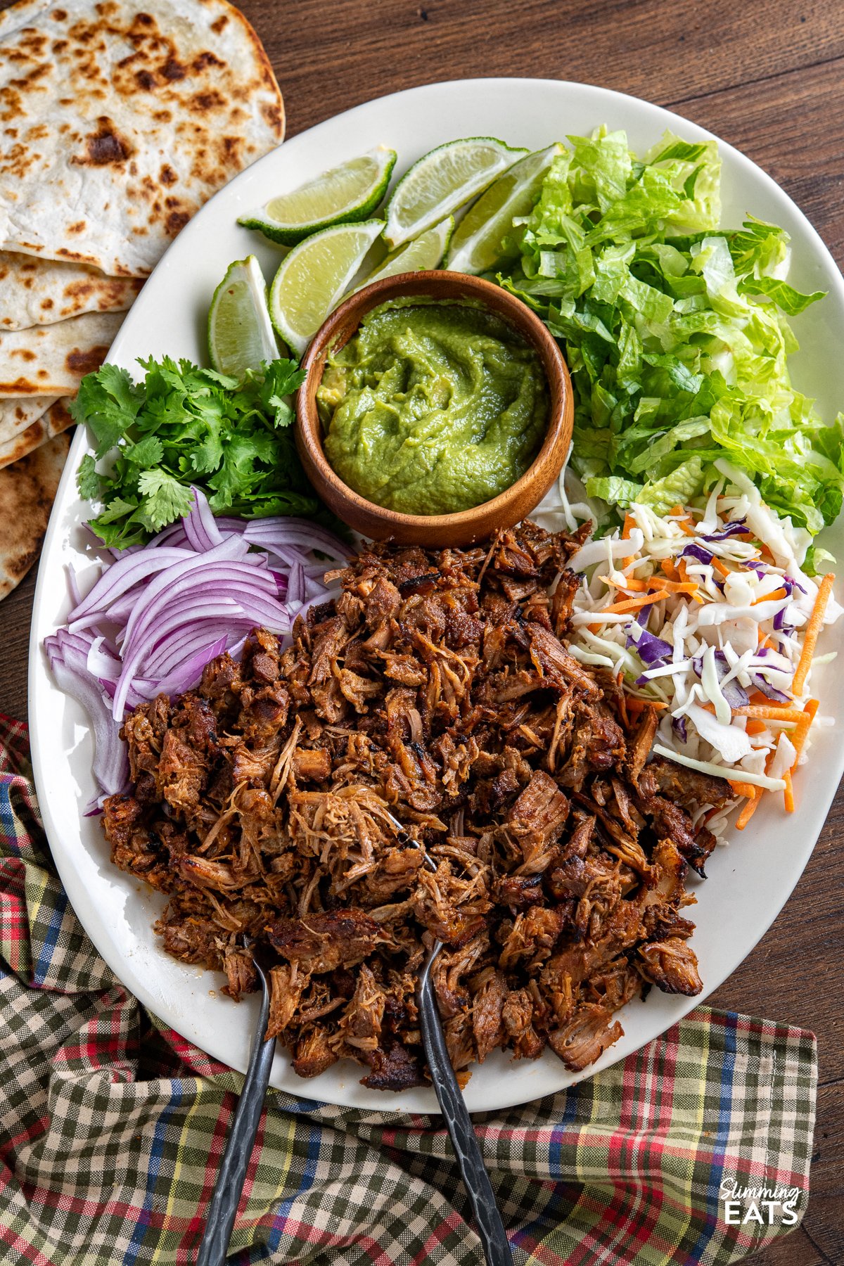 Mexican Pineapple Pork Carnitas on large oval plate with red onion, slaw, lettuce, cilantro, wooden bowl of guacamole and lime wedges