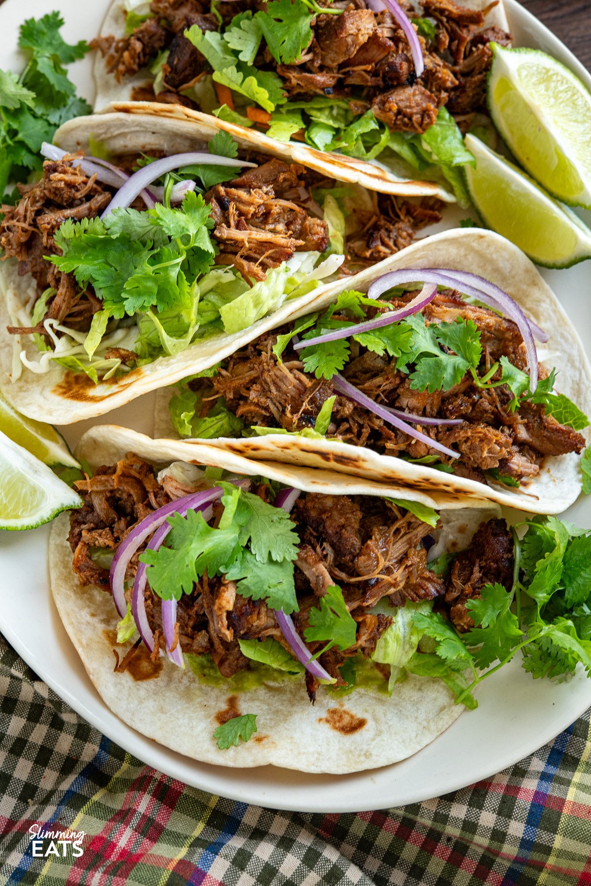 Get ready to tantalize your taste buds with these mouthwatering Pineapple Mexican Pork Carnitas, a flavourful twist on traditional pork carnitas that will become your new favourite pork recipe and can be perfectly cooked in a slow cooker or pressure cooker. close up of Mexican Pineapple Pork Carnitas in soft tortillas topped with cilantro, red onion and guacamole with lime wedges on side