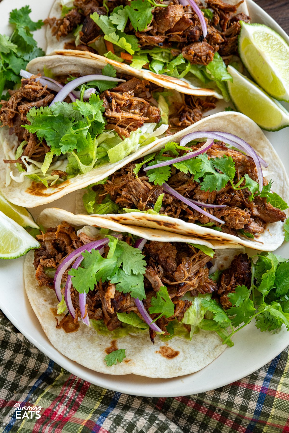 close up of Mexican Pineapple Pork Carnitas in soft tortillas topped with cilantro, red onion and guacamole with lime wedges on side