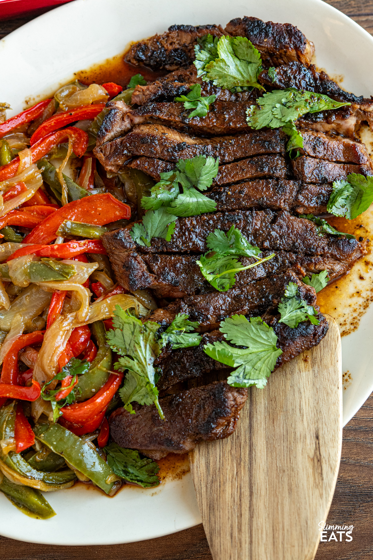 Tender, flavour packed steak marinated in spices and finished with a sweet savory glaze for a simple yet delicious meal. close up of Marinated Mouth Watering Steak on oval cream plate with sauteed onions and peppers, red dish with side of potatoes visible above.